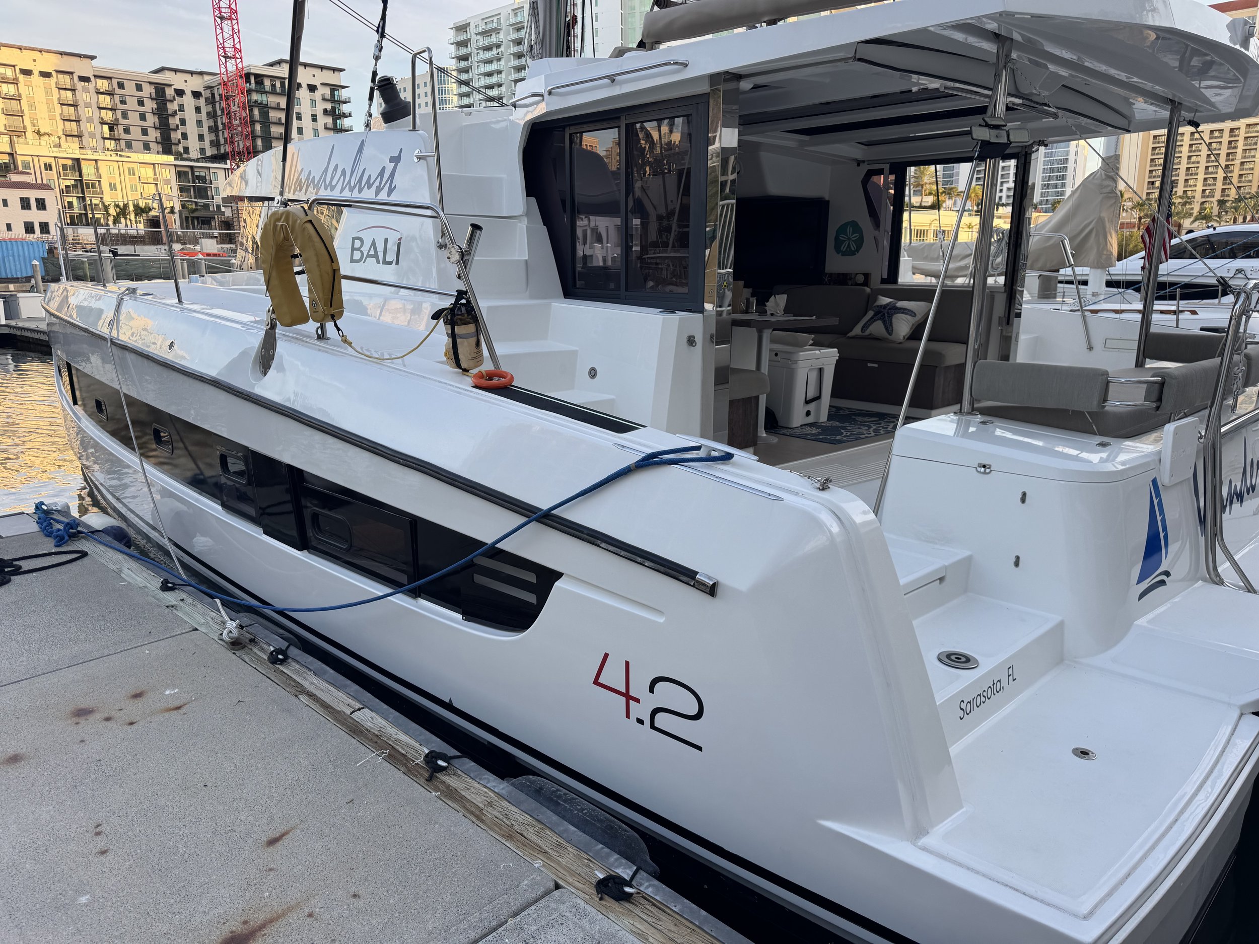 Catamaran docked in a marina with "Wanderlust" and "Bali 4.2" on its side, featuring an open deck and modern interior, with surrounding buildings.