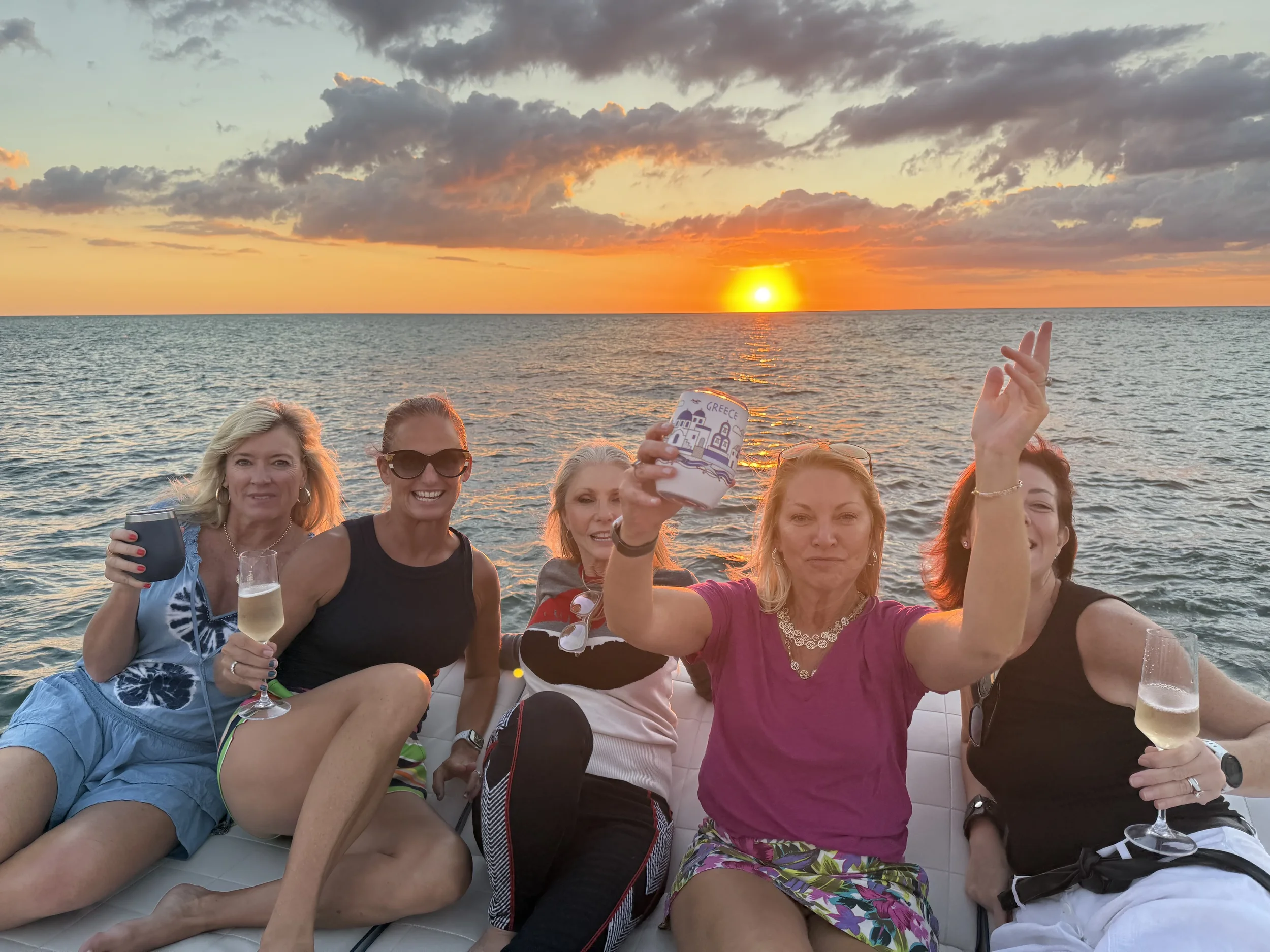 A group of five women celebrating on a boat during sunset, holding drinks and smiling.