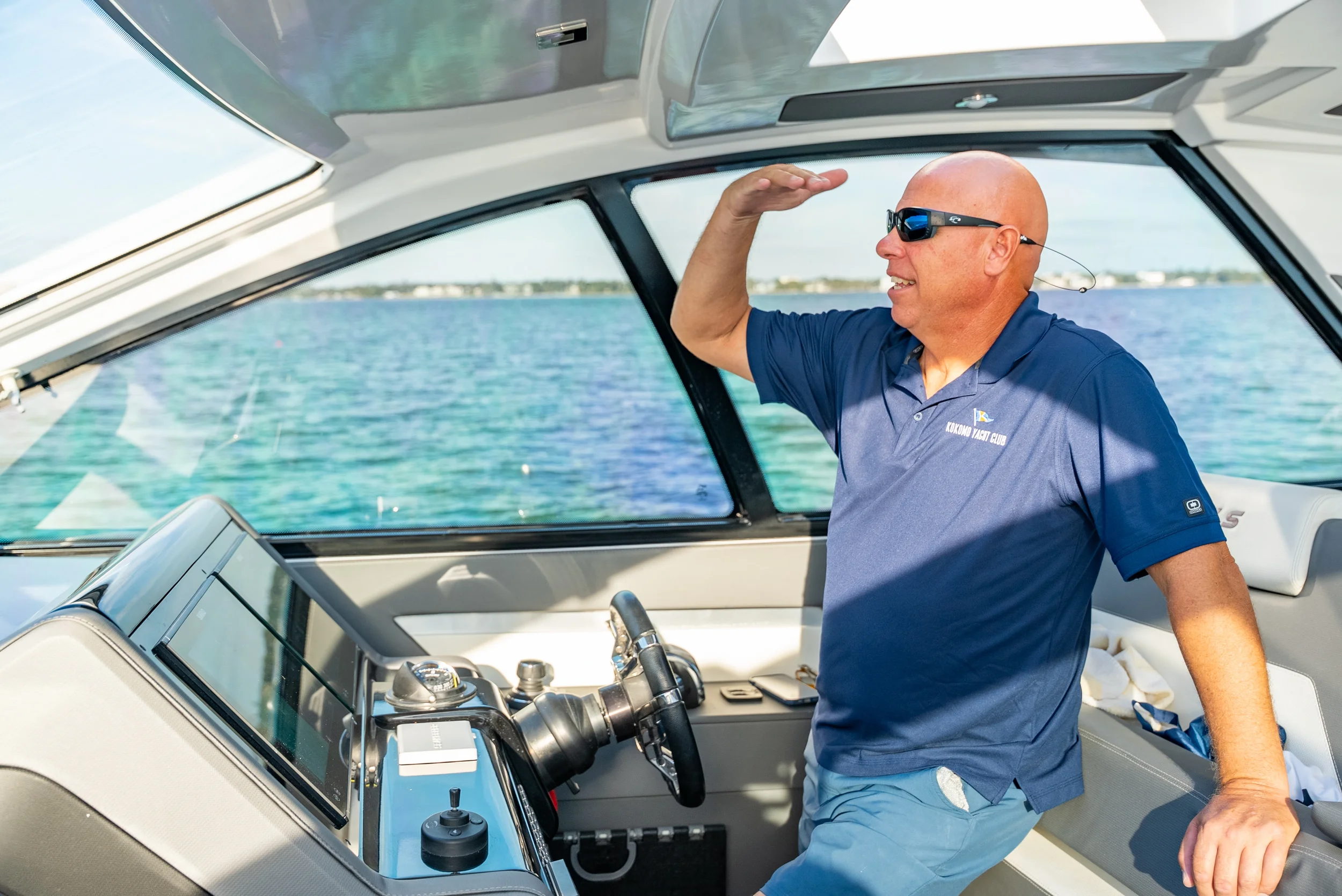 Kokomo charters - luxury yacht charter -A man on a boat, wearing sunglasses and a navy polo shirt, is saluting with his right hand while holding onto the boat's console with his left. The water and coastline are visible through the boat's windshield.