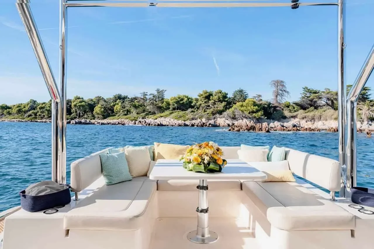 kokomo charters - 50' Memory Maker - View of the back deck of a boat with white cushioned seating, a central table with a yellow flower arrangement, and a scenic water and tree-lined shoreline in the background.