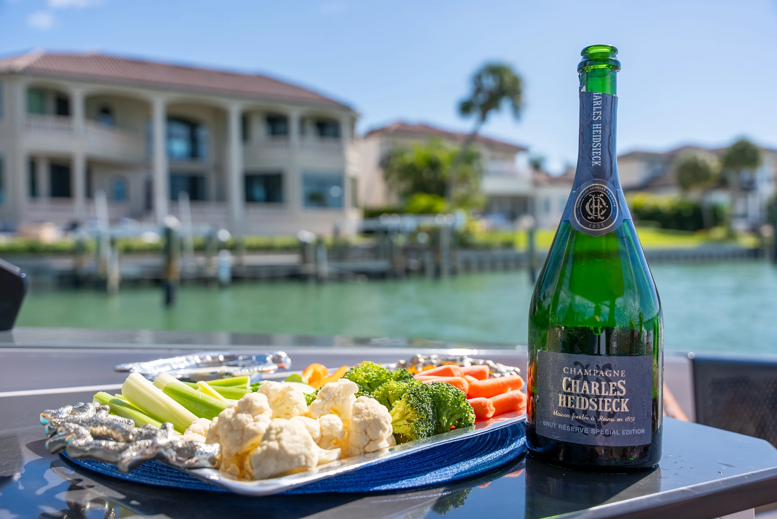 Kokomo charters - yacht float plan Sarasota - SA bottle of Charles Heidsieck champagne and a platter of assorted fresh vegetables, including cauliflower, celery, broccoli, and carrots, on an outdoor table near a waterway.