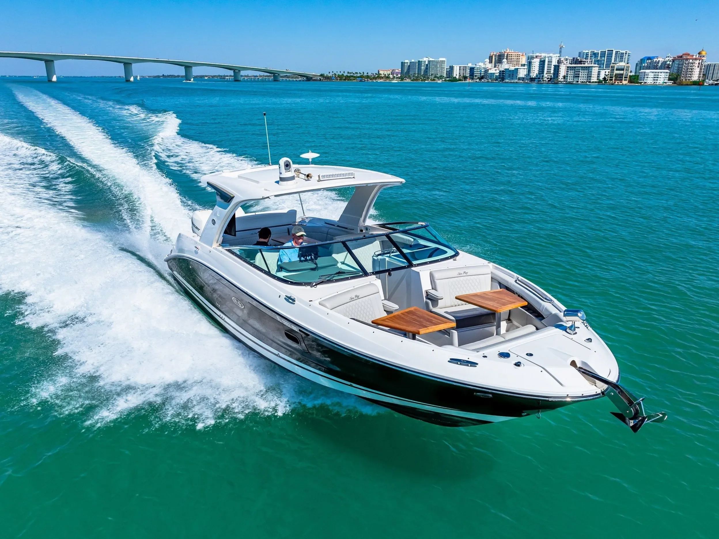 A white motorboat speeding through turquoise water near a city skyline with high-rise buildings.