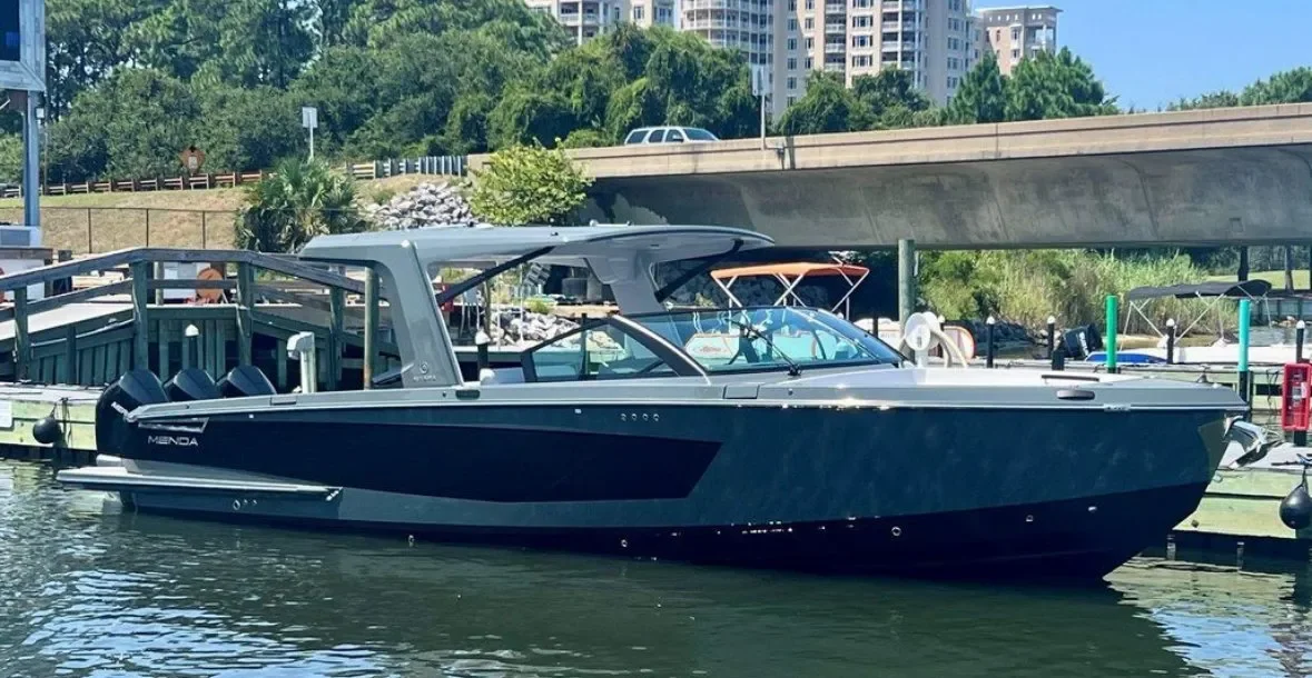 A sleek, black luxury motor yacht named Menda docked at a marina with trees, buildings, and a bridge in the background.