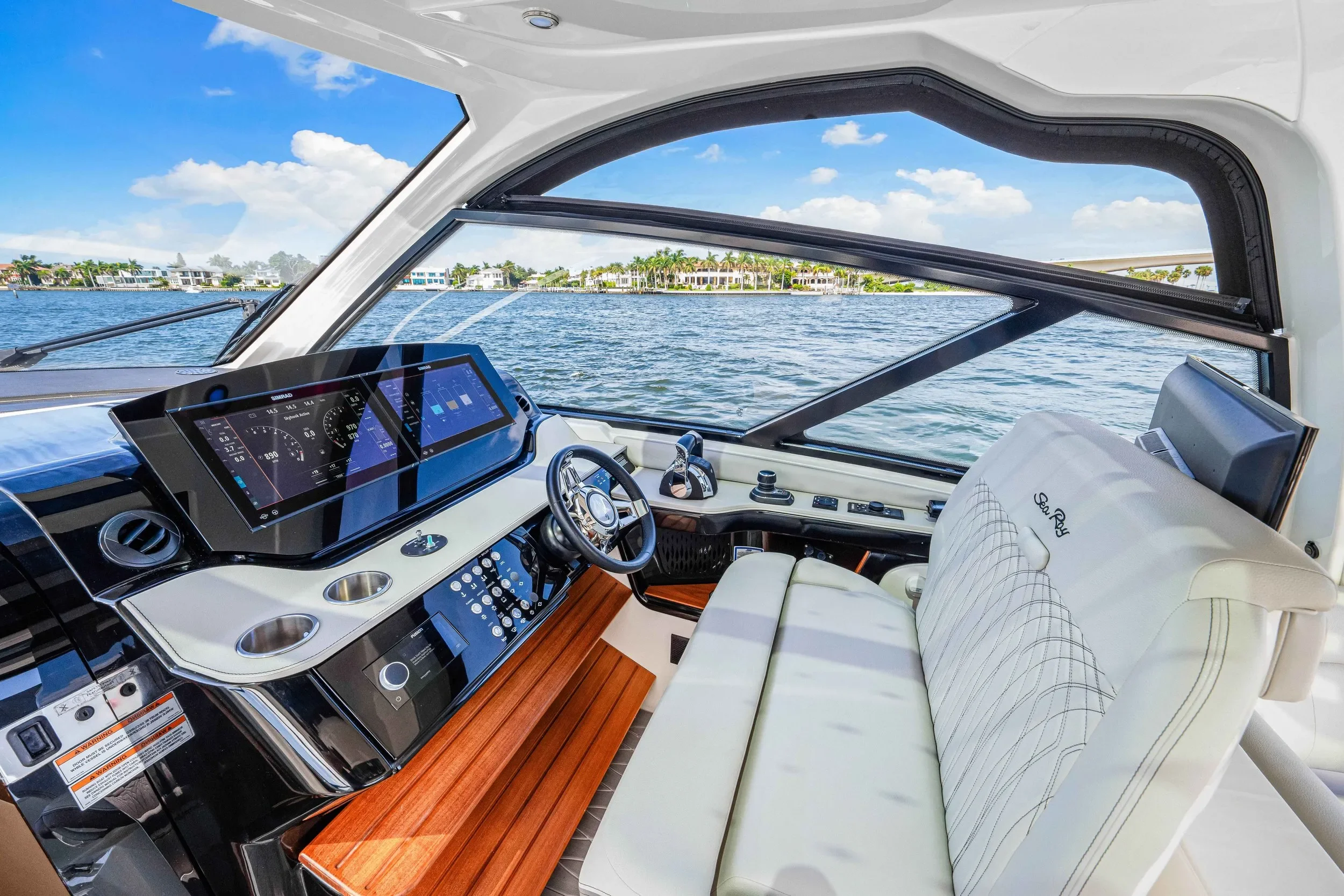 kokomo charters - 40' Golden Ticket - Interior view of a luxury yacht's helm station with a large digital dashboard, steering wheel, and seating, overlooking water and a distant shoreline with houses and palm trees under a partly cloudy sky.