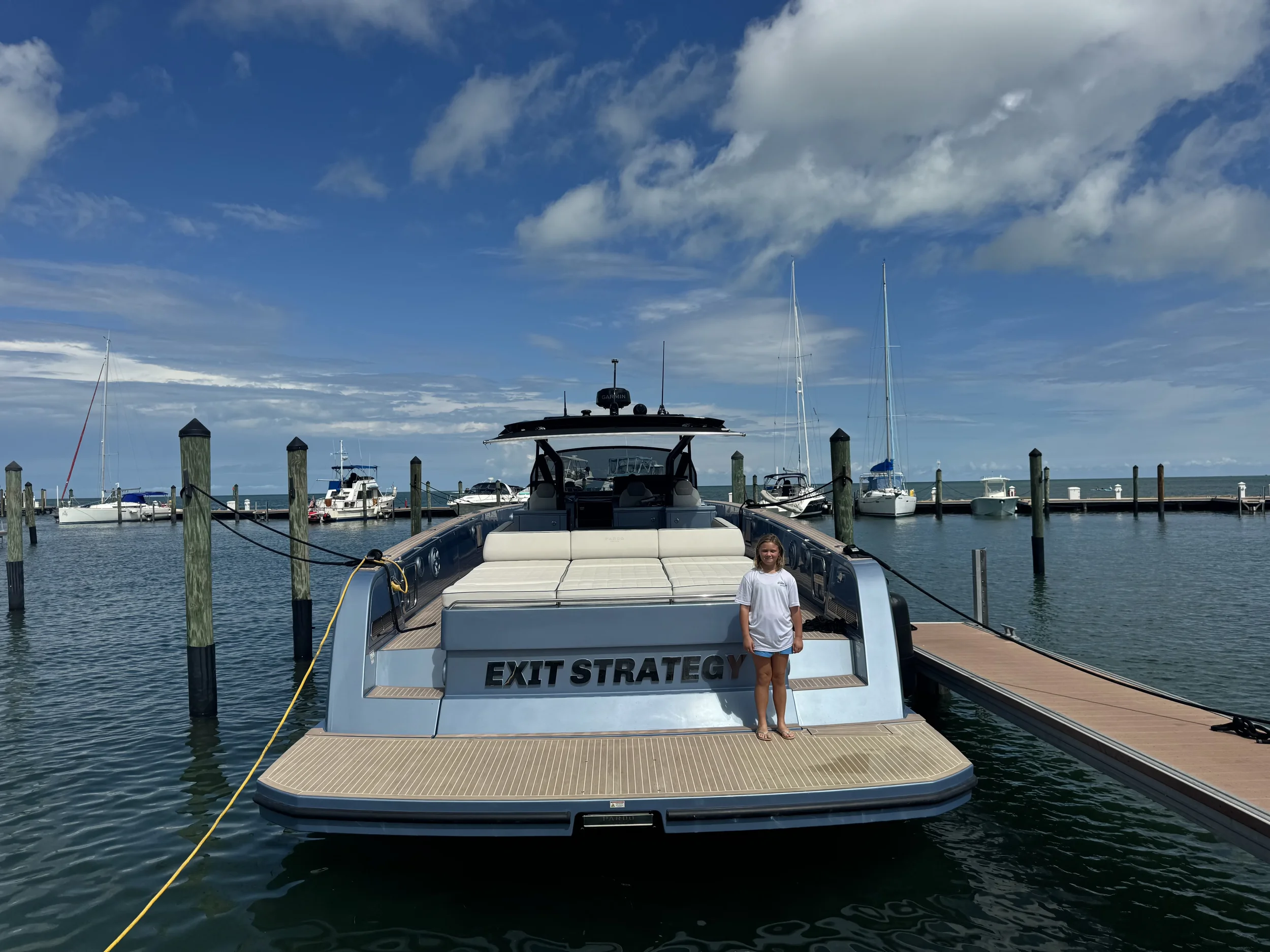 A woman in a white t-shirt and shorts standing on the back of a luxury yacht named 'EXIT STRATEGY' docked at a marina with other boats in the background under a blue sky with clouds.