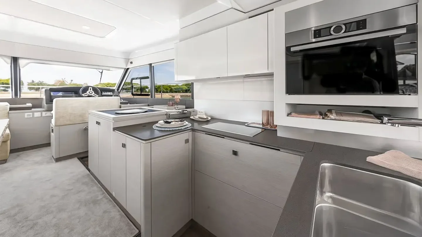 Interior of a modern yacht kitchen with white cabinets, black countertops, and stainless steel appliances, with large windows showing an outdoor view.