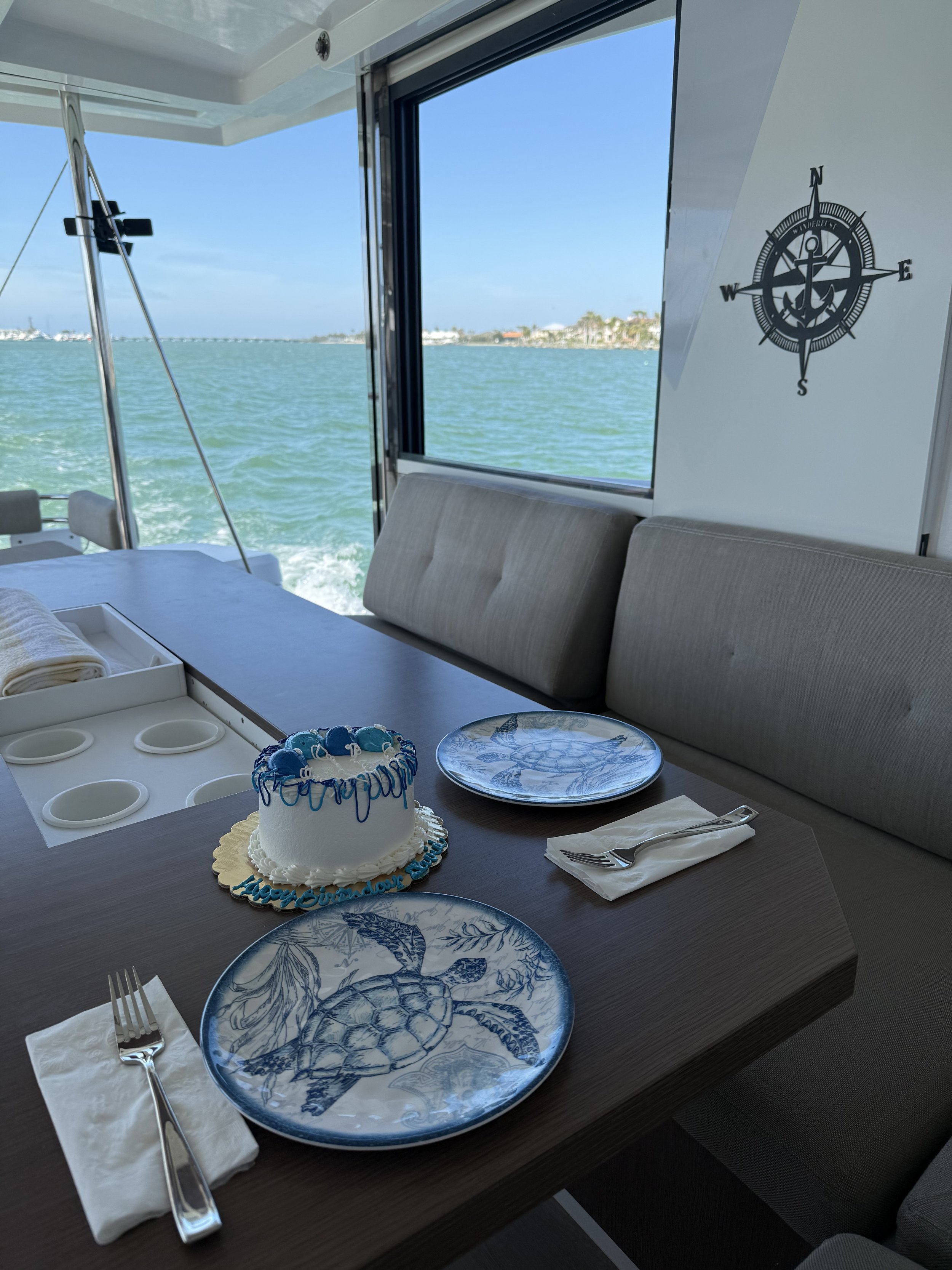 Interior of a boat with a table set for two, featuring a small cake and two sea turtle-themed plates. A compass design is visible on the wall, with an ocean view through the window.