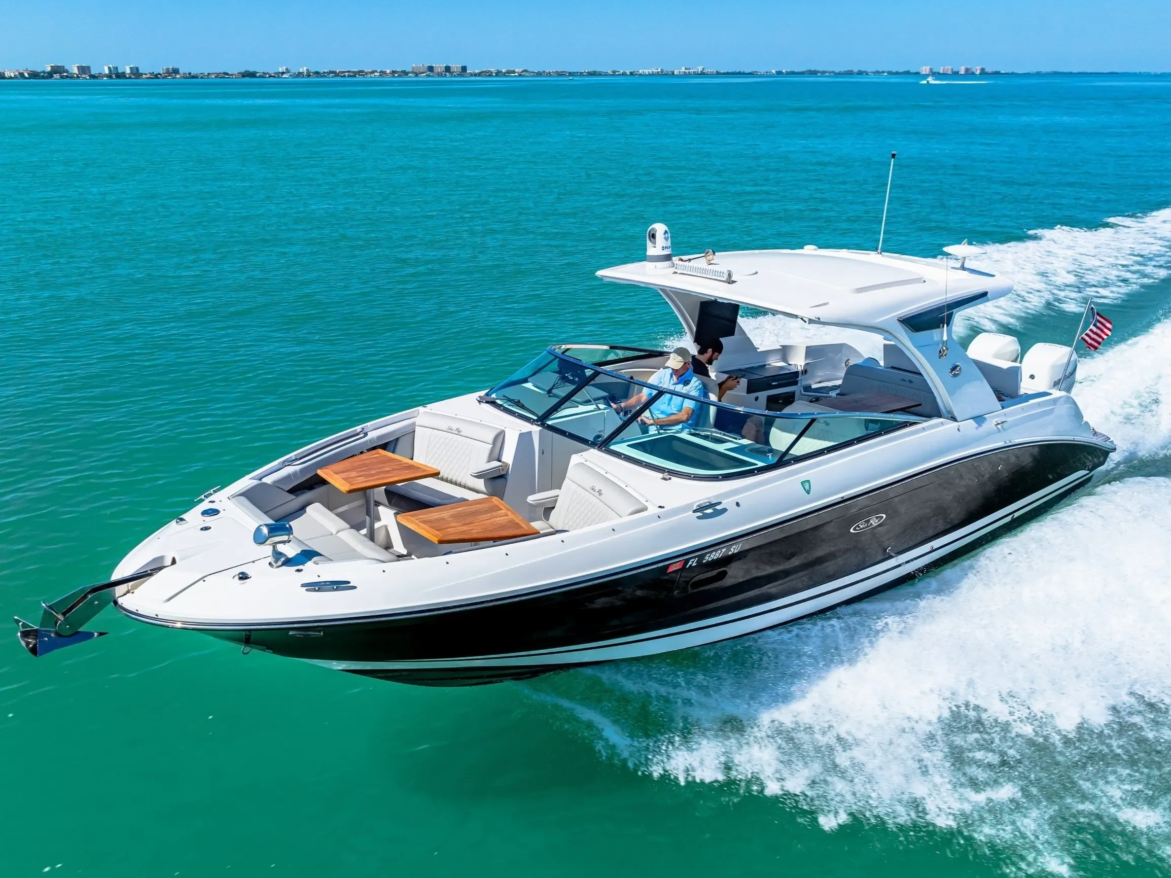 A white and black motor yacht speeding across turquoise water with a city skyline in the background, displaying a spacious deck with wooden tables and seating.