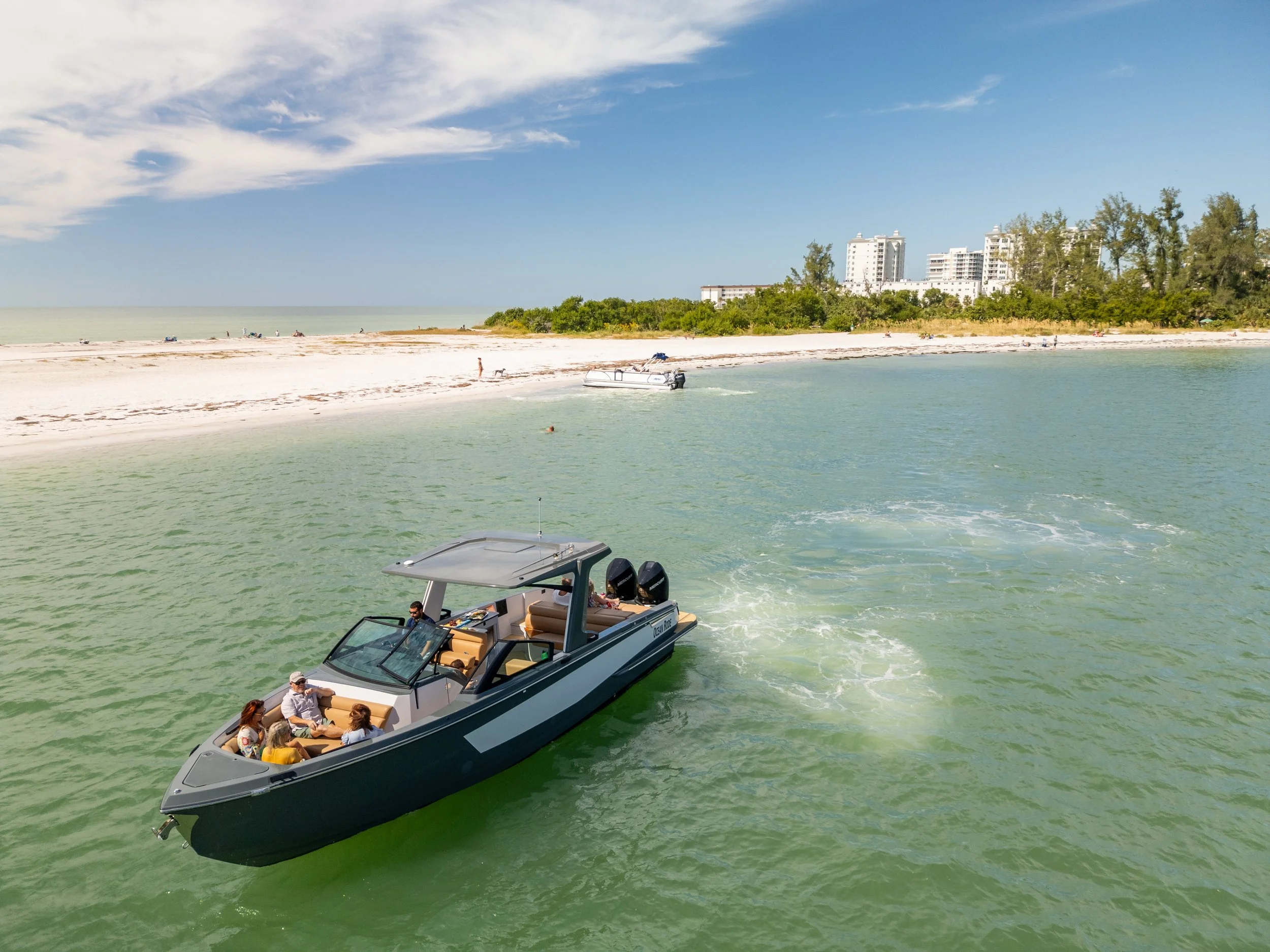 Kokomo charters - yacht float plan Sarasota - A group of people riding on a black and white motorboat near a sandy beach with white buildings and green trees in the background.