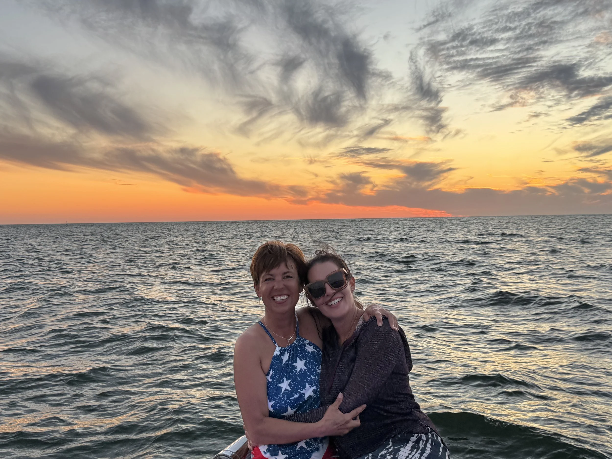 Two smiling women embracing on a boat with ocean and sunset.