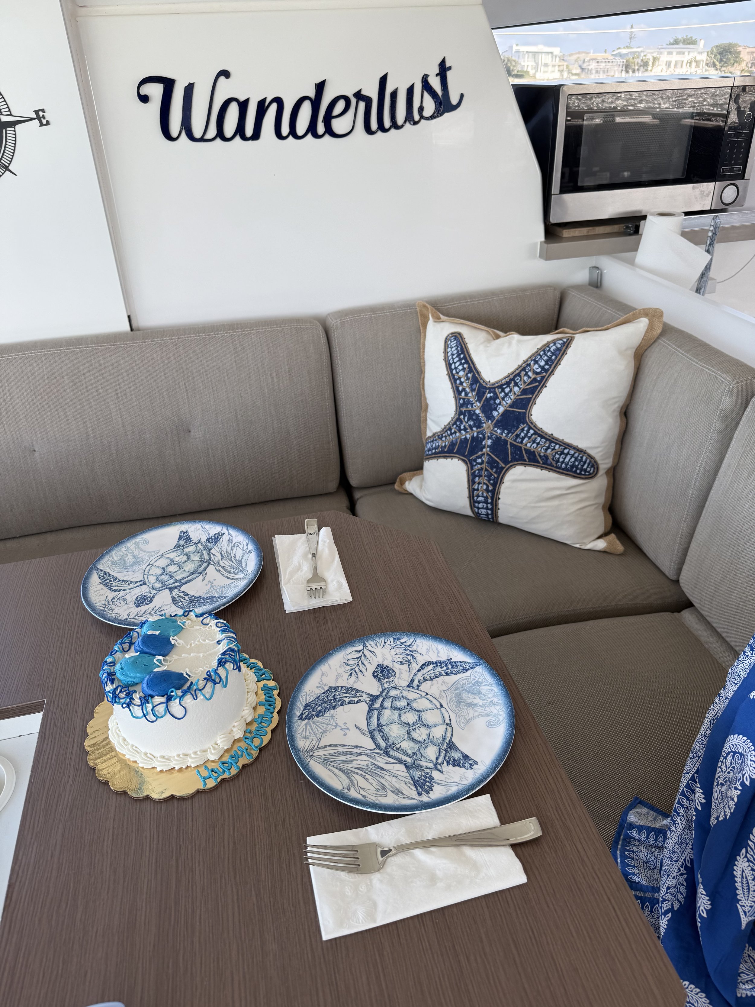 Interior of a room with a couch featuring a starfish pillow, a wall sign reading 'Wanderlust,' and a table set with two turtle-themed plates, forks on napkins, and a small cake with blue frosting and decorations next to a microwave.