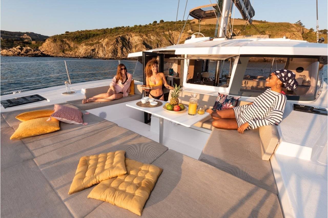 Three people relaxing on a catamaran yacht with cushions, fruit, and drinks on a table, near a rocky coastline.