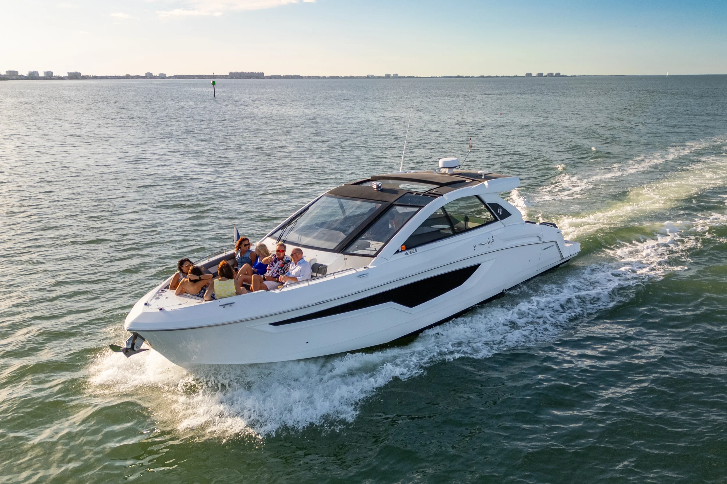 Kokomo charters - bachelorette boat party Sarasota - A white motor yacht sailing on the water with six people on the deck, some wearing sunglasses and summer clothes, under a clear sky with distant shoreline and buildings in the background.
