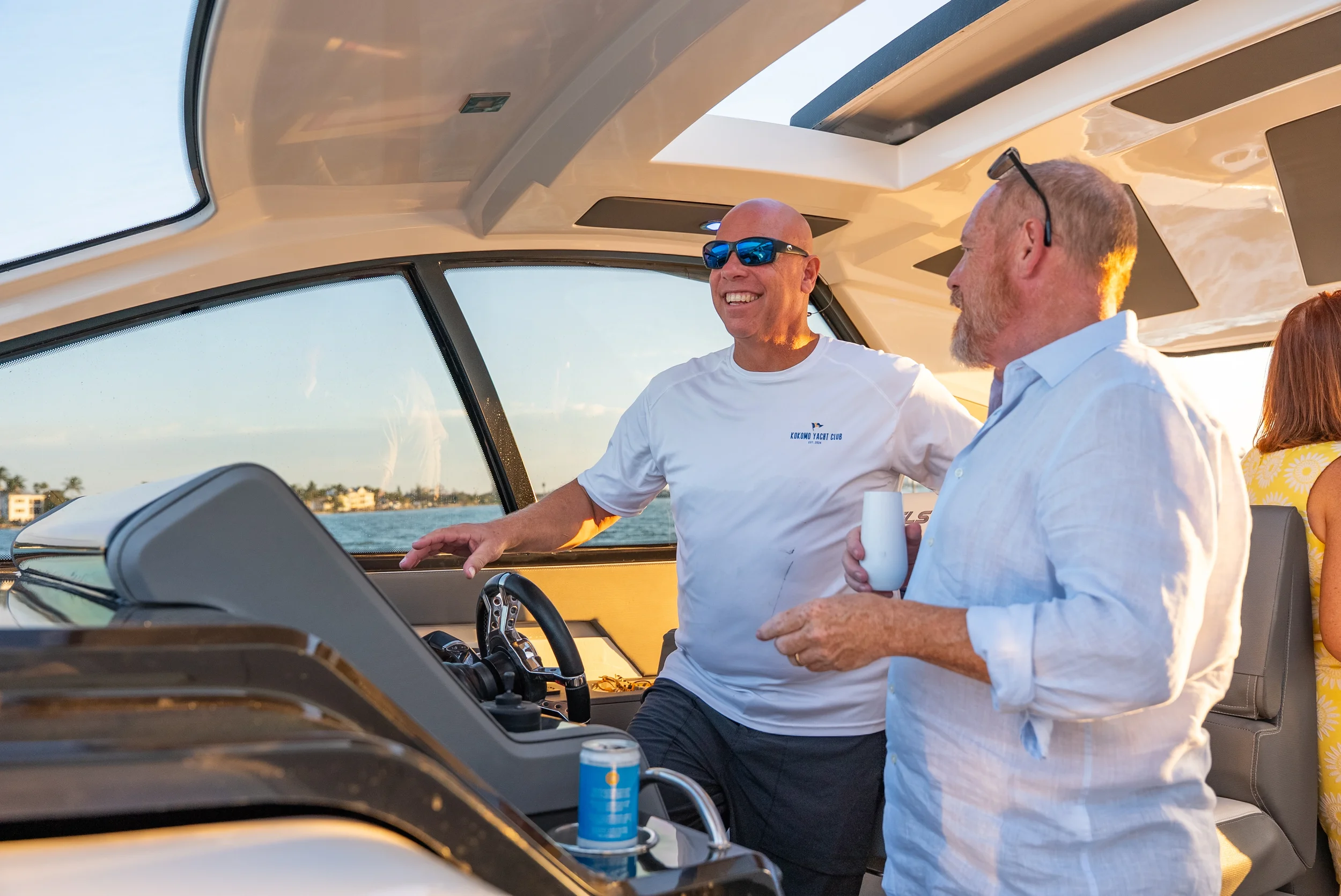 Kokomo charters - Sarasota private yacht charter - Two men smiling and chatting on a yacht during sunset, one wearing sunglasses and a white t-shirt, the other in a white shirt holding a drink.