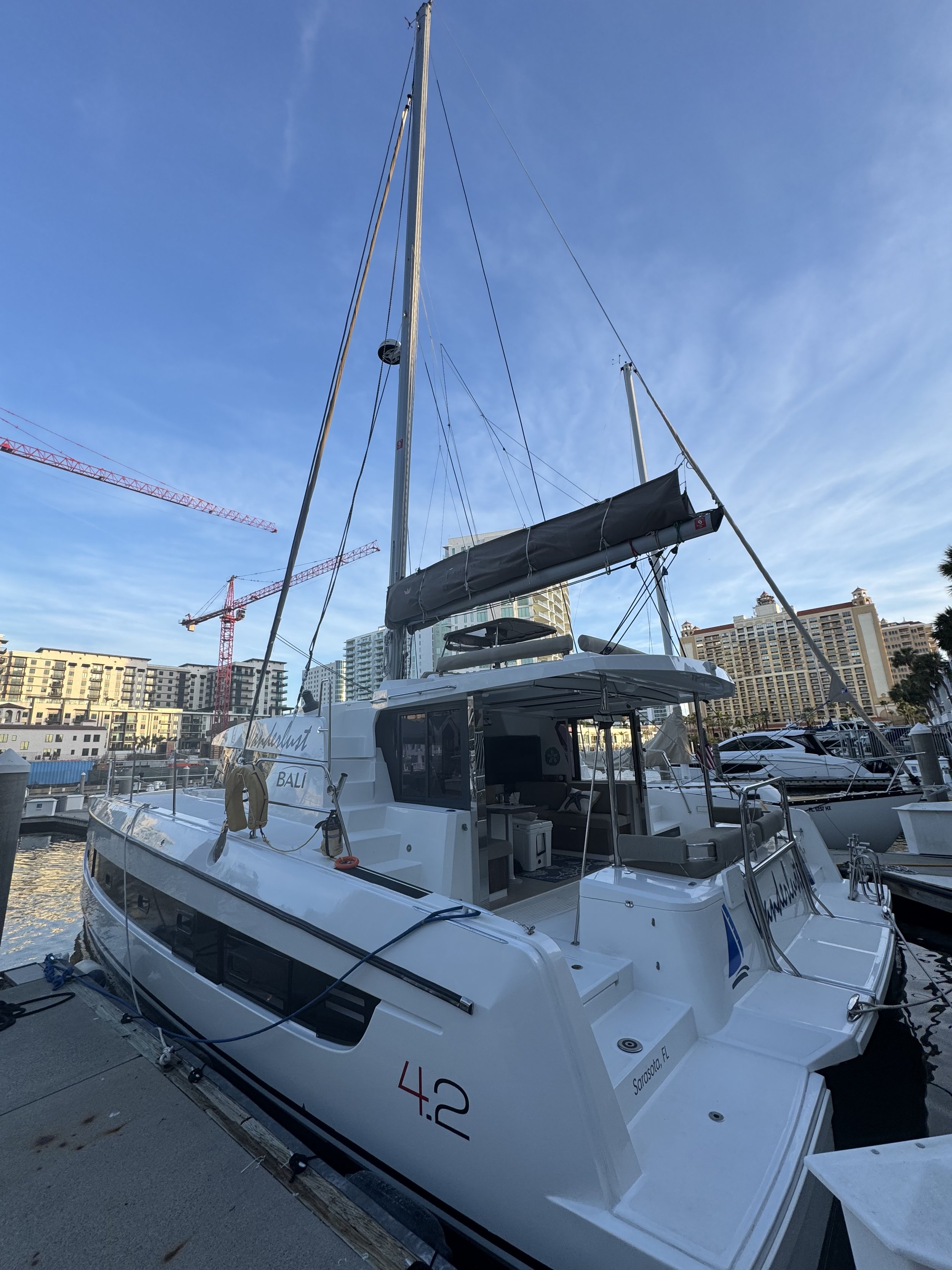 A white catamaran with the number 4.2 on its side, docked in a marina with modern buildings and cranes in the background.