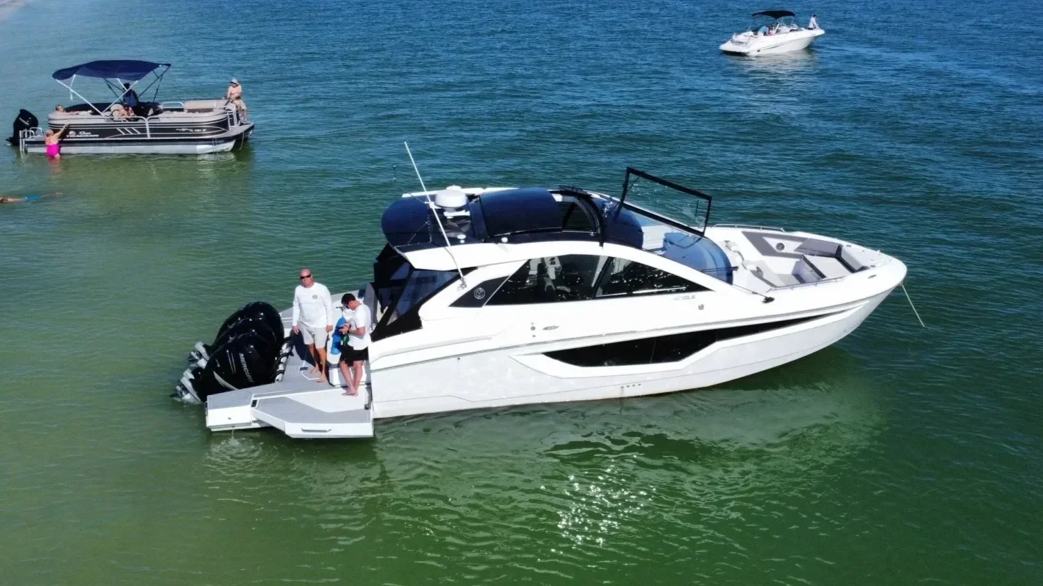 A large white yacht with a black top docked in the water, with three people on deck, and two people standing nearby. Several smaller boats are visible in the background on the water.