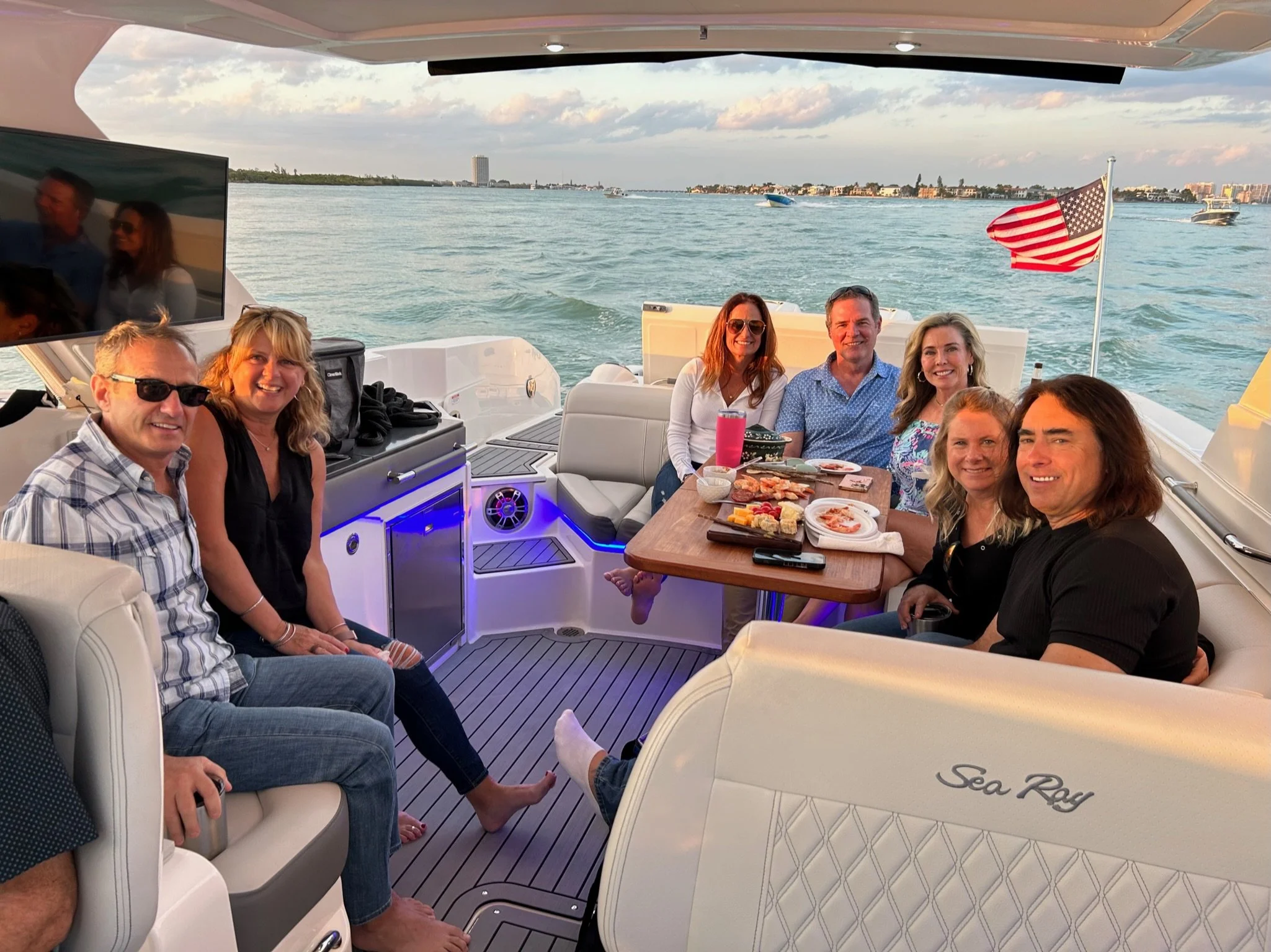 Group of people enjoying a gathering on a Sea Ray boat, with food on a table and an American flag at the stern; water and shoreline visible in the background.
