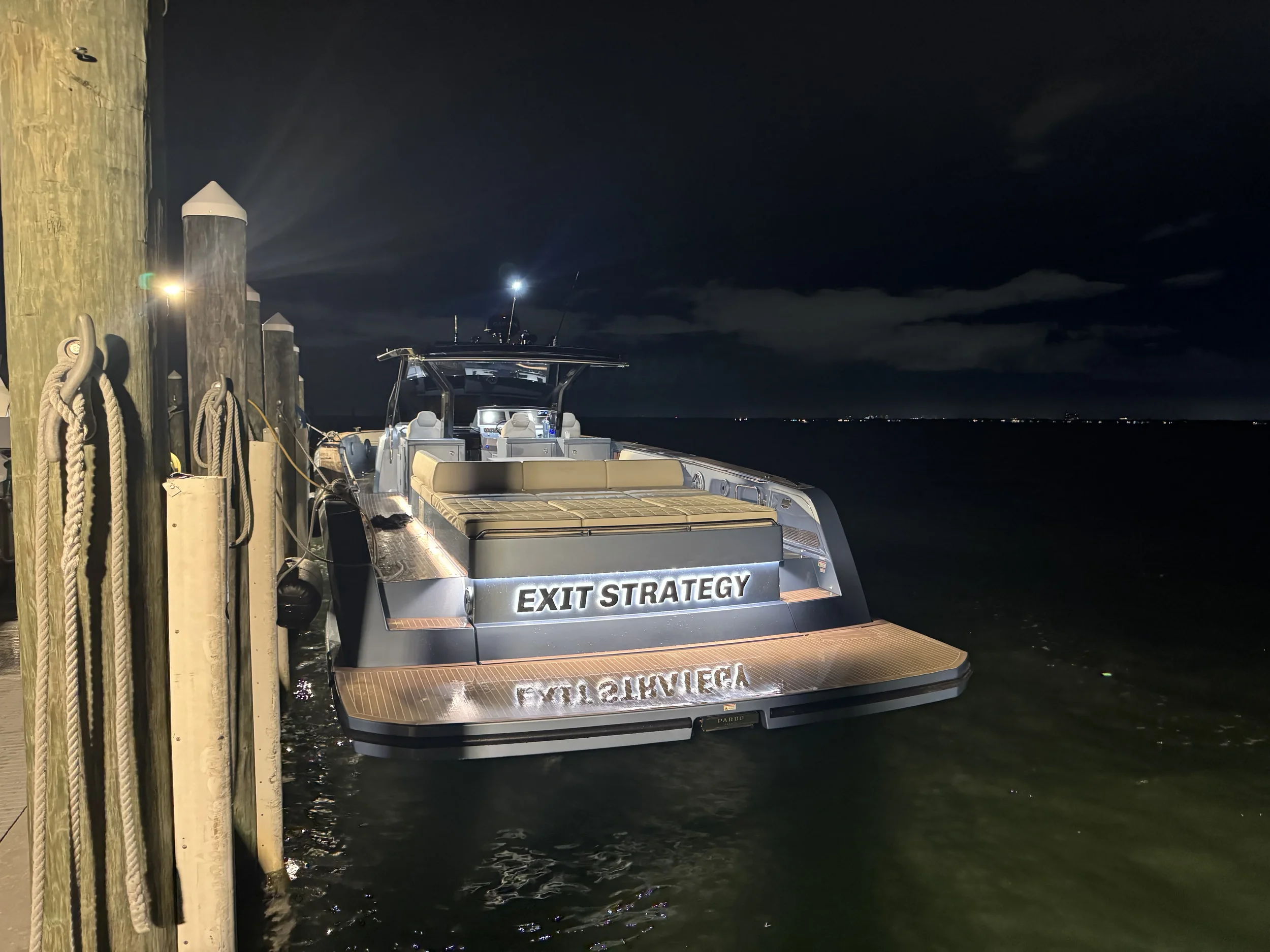 A modern boat docked at a wooden pier at night with a sign that reads 'EXIT STRATEGY' on the back.