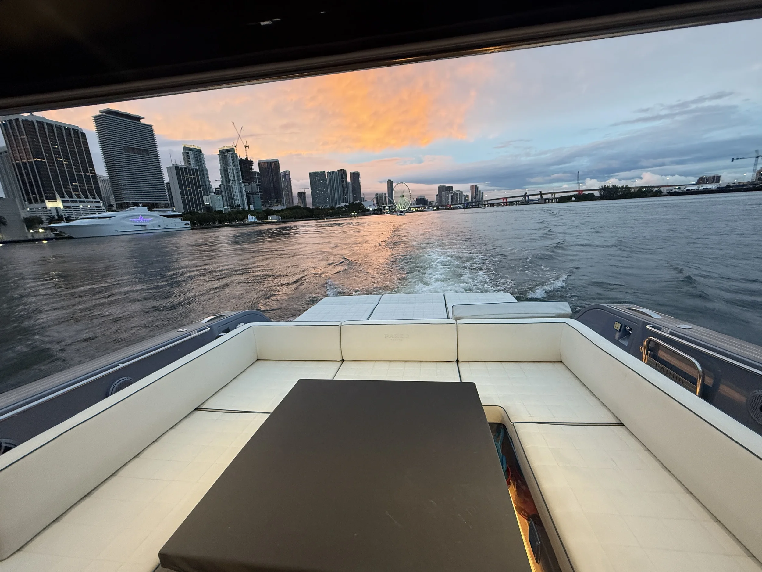 View from the back of a boat on a body of water, overlooking a city skyline at sunset with tall buildings, cranes, and a ferris wheel.