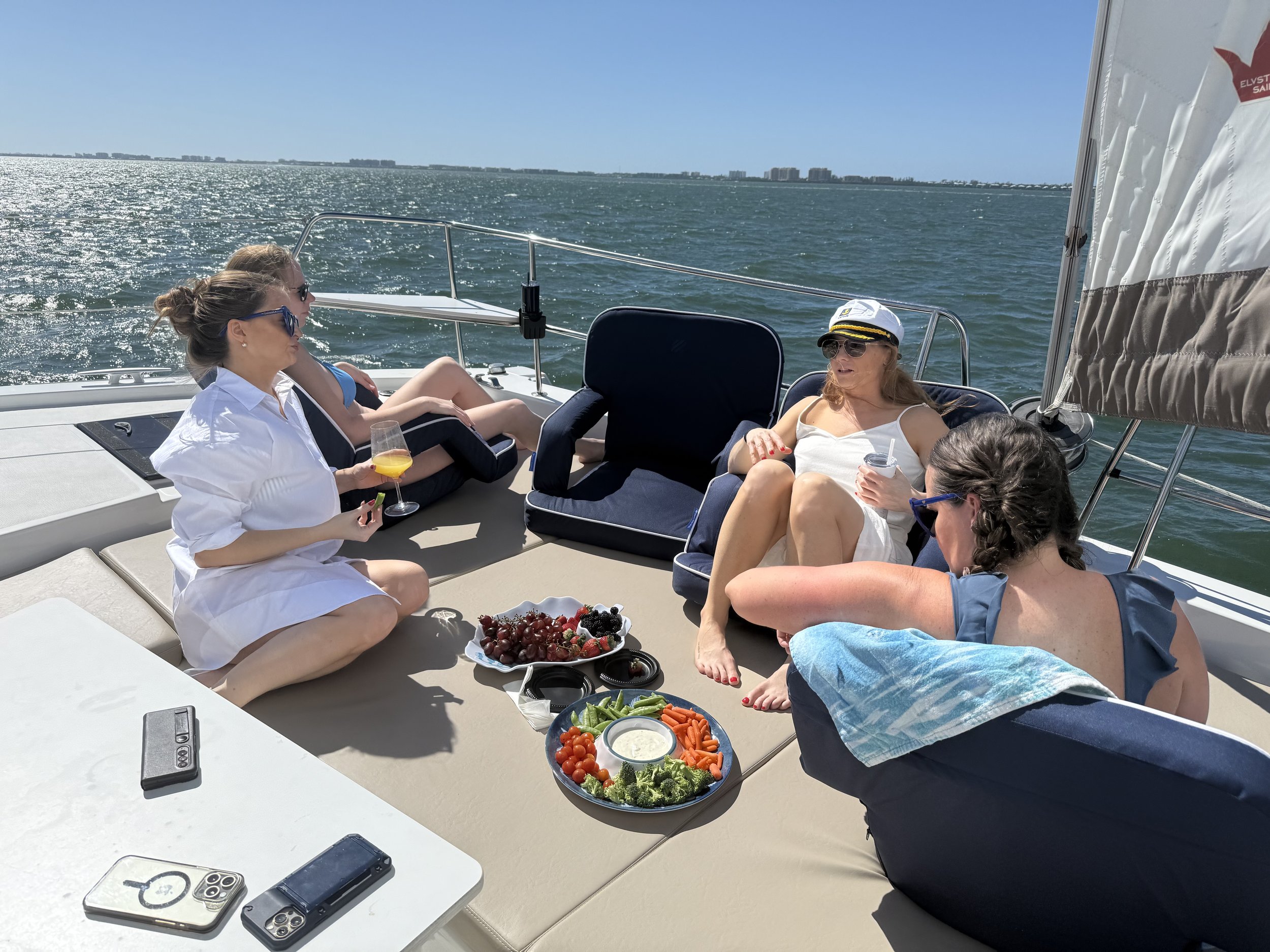 Four women relaxing on a boat deck with snacks and drinks, near a sunny waterfront.