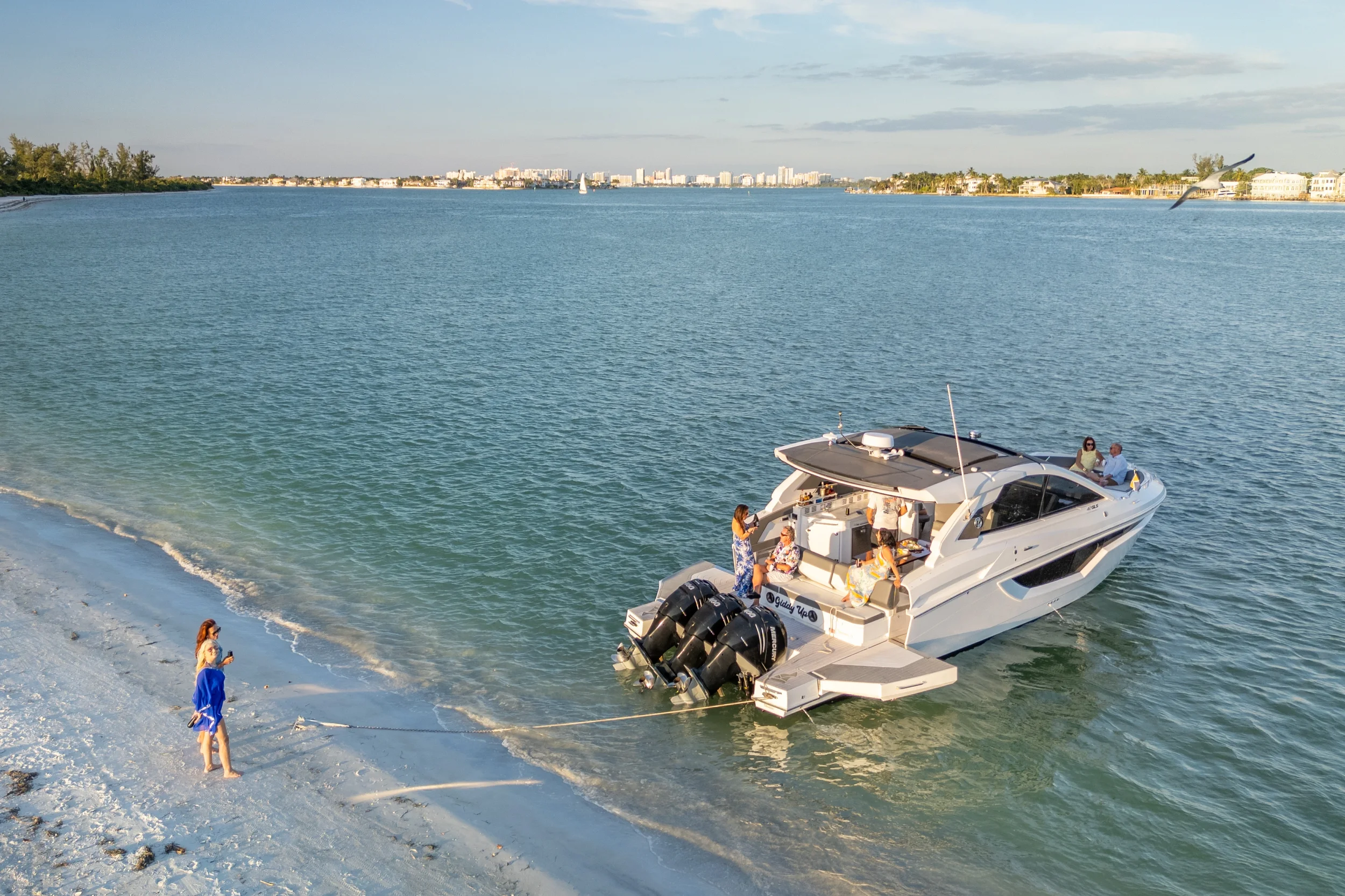 Kokomo charters - Sarasota private yacht charter - A white motor yacht docked near a sandy beach with seven people on and around it, some sitting on the boat and others on the sand, with a woman in a blue dress standing on the beach holding a phone.