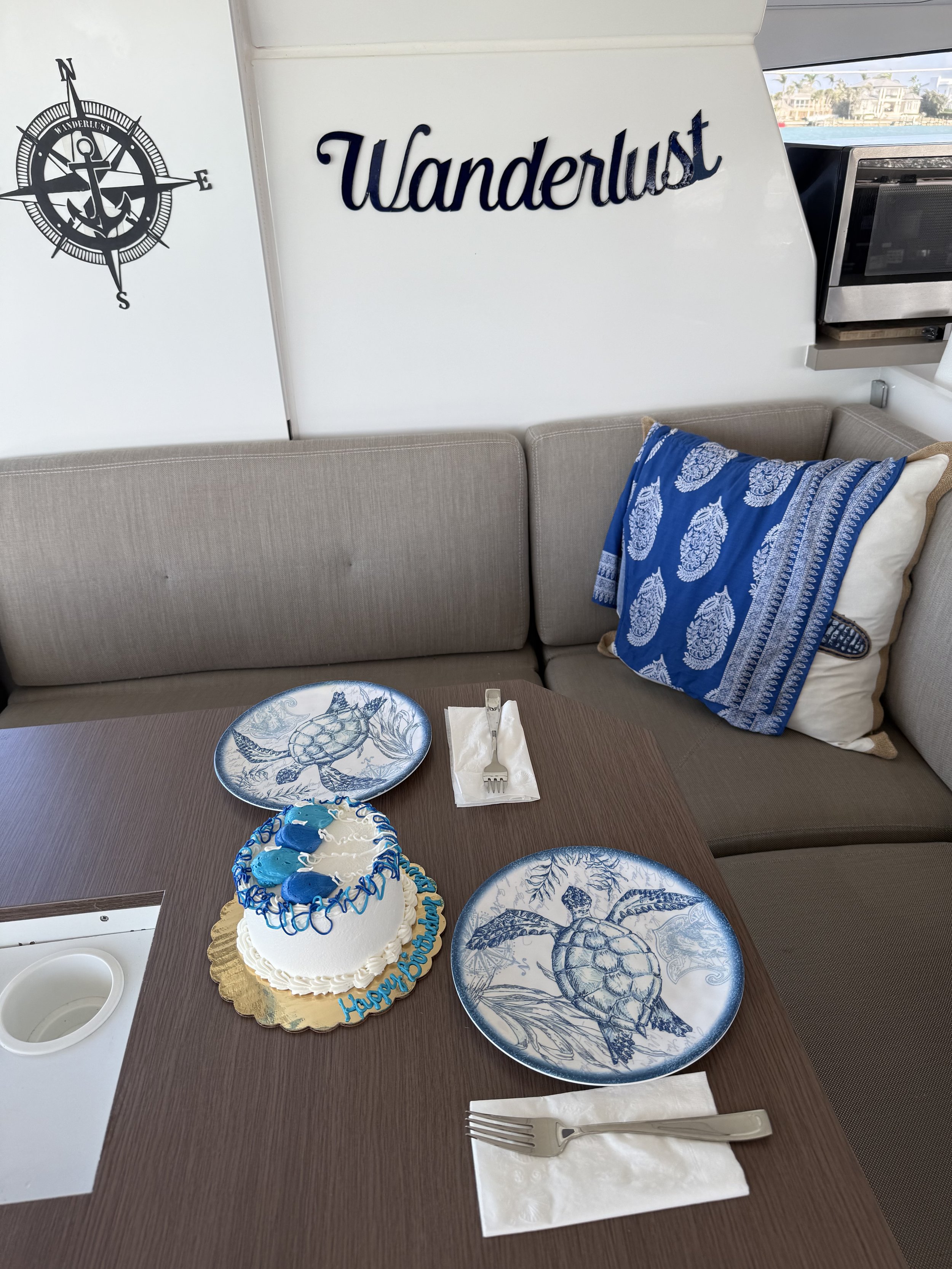 Dining table on a boat with 'Wanderlust' sign, two plates featuring sea turtle designs, a small cake with a blue and white nautical theme, and a compass wall decal. Forks and napkins are placed next to each plate. A cushion with marine prints is on t