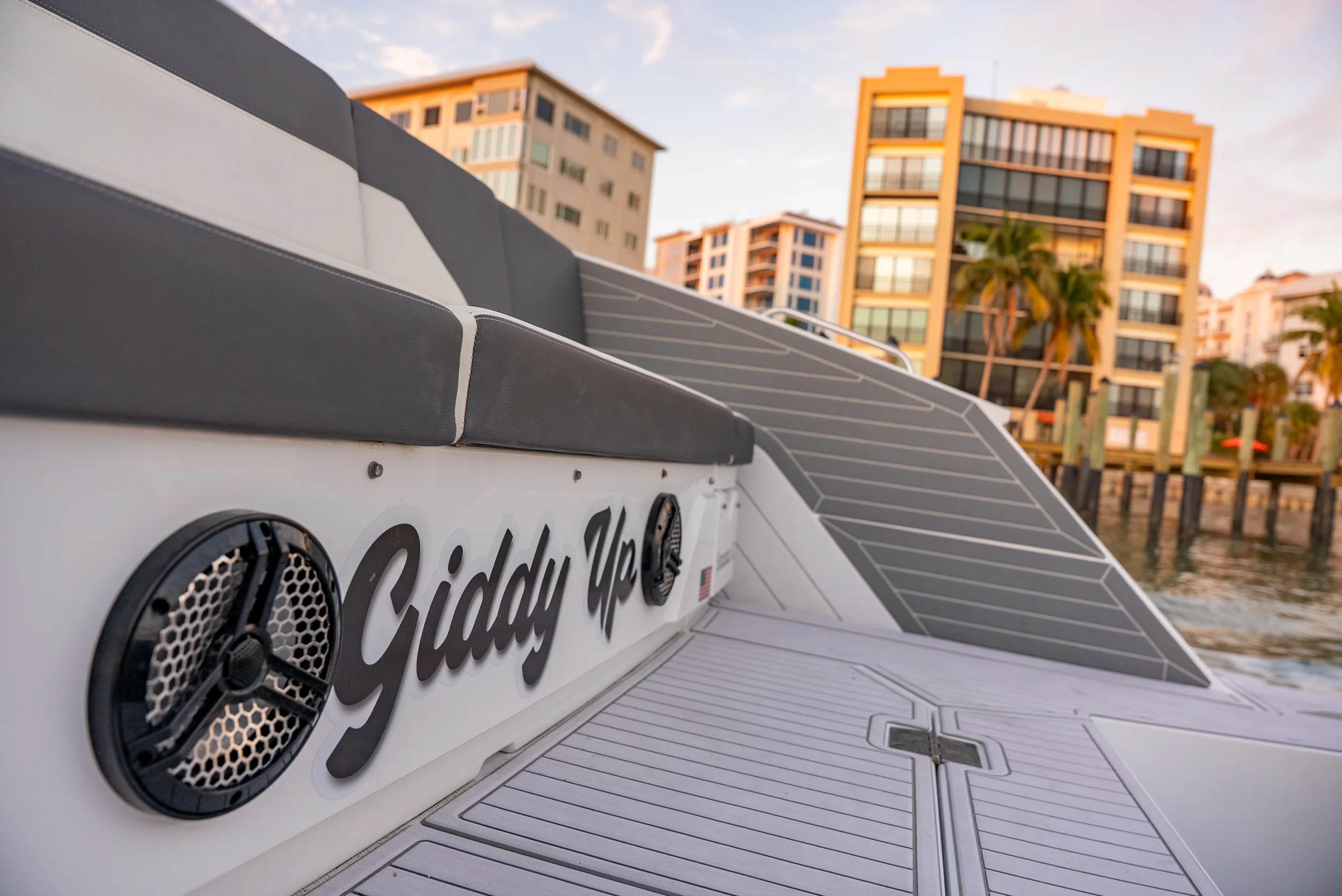 Kokomo charters - St. Pete sunset cruise -  Close-up of a boat's starboard side with the name 'Giddy Up' on the hull, showing speakers and seating, with modern apartment buildings and palm trees in the background.