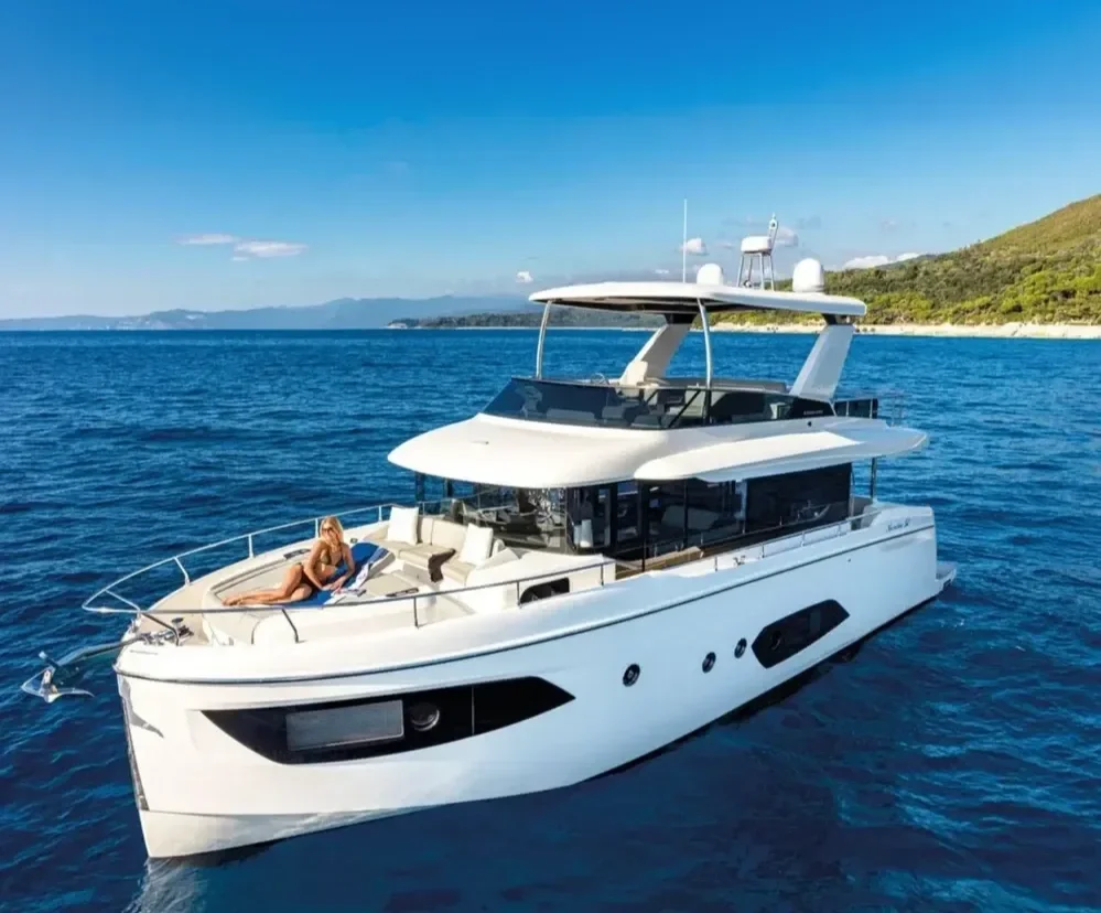 A luxury white yacht floating on calm blue ocean water with a woman sunbathing on the deck, and a distant shoreline with hills in the background under a clear blue sky.