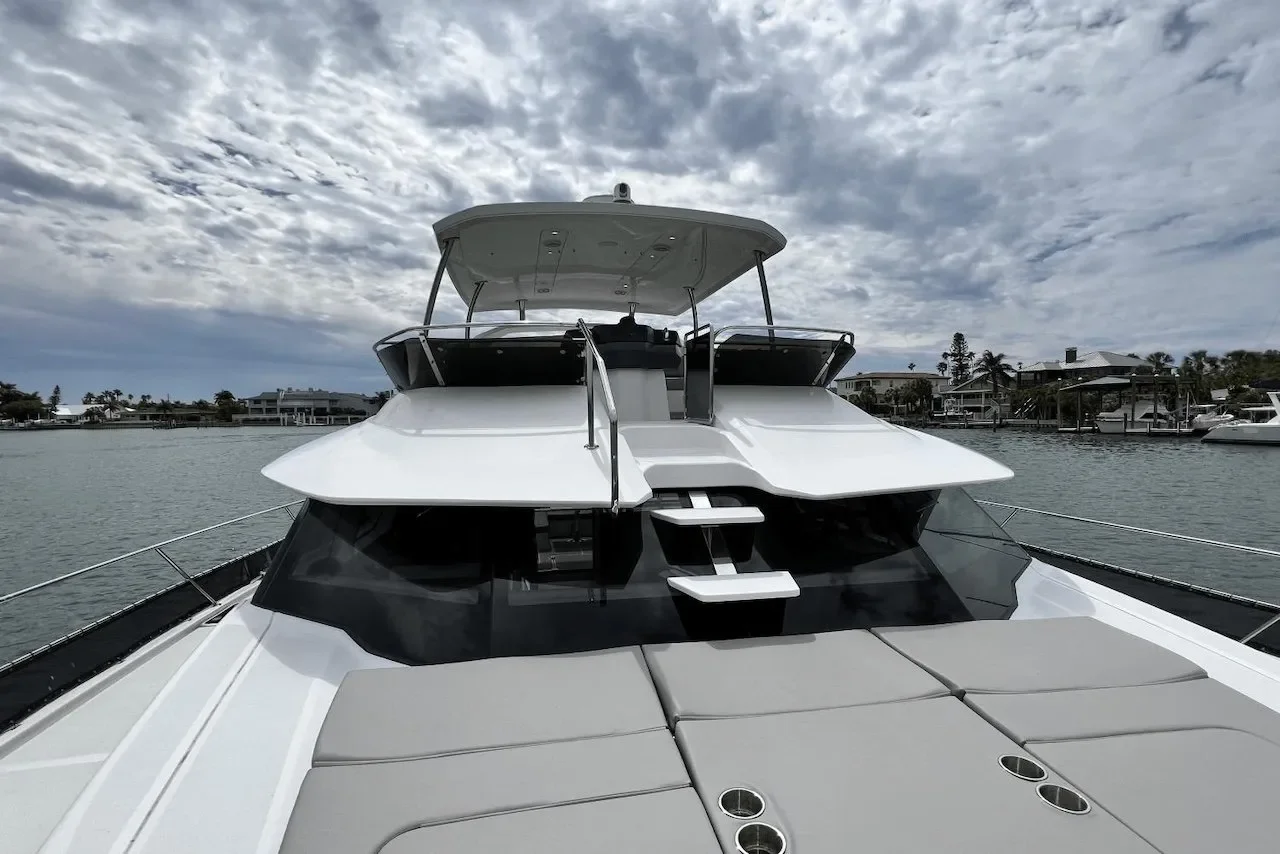 Front view of a modern white yacht anchored in a waterway with homes and trees in the background, under a cloudy sky.