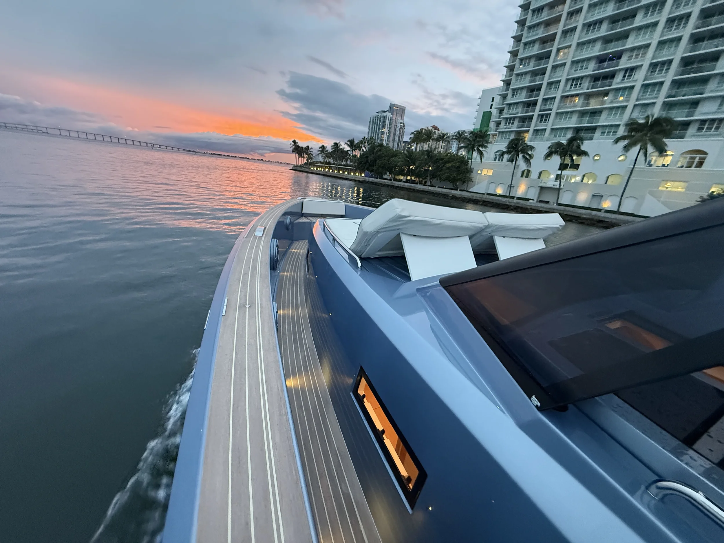 View from a yacht on the water at sunset, with modern buildings and palm trees along the shoreline in the background.