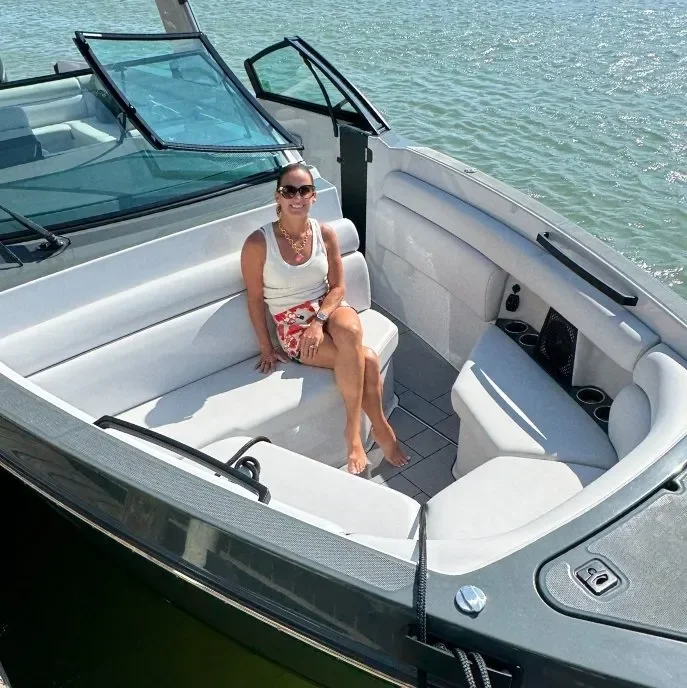Woman sitting on white cushioned bench inside a boat, wearing sunglasses, a white top, red patterned shorts, and jewelry, on a sunny day with water in the background.