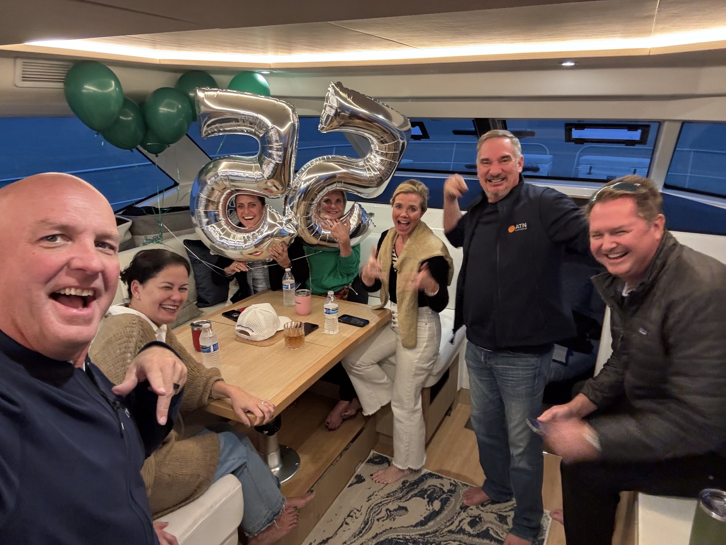Group of six people on a boat celebrating a 55th birthday with silver balloons and green decorations.
