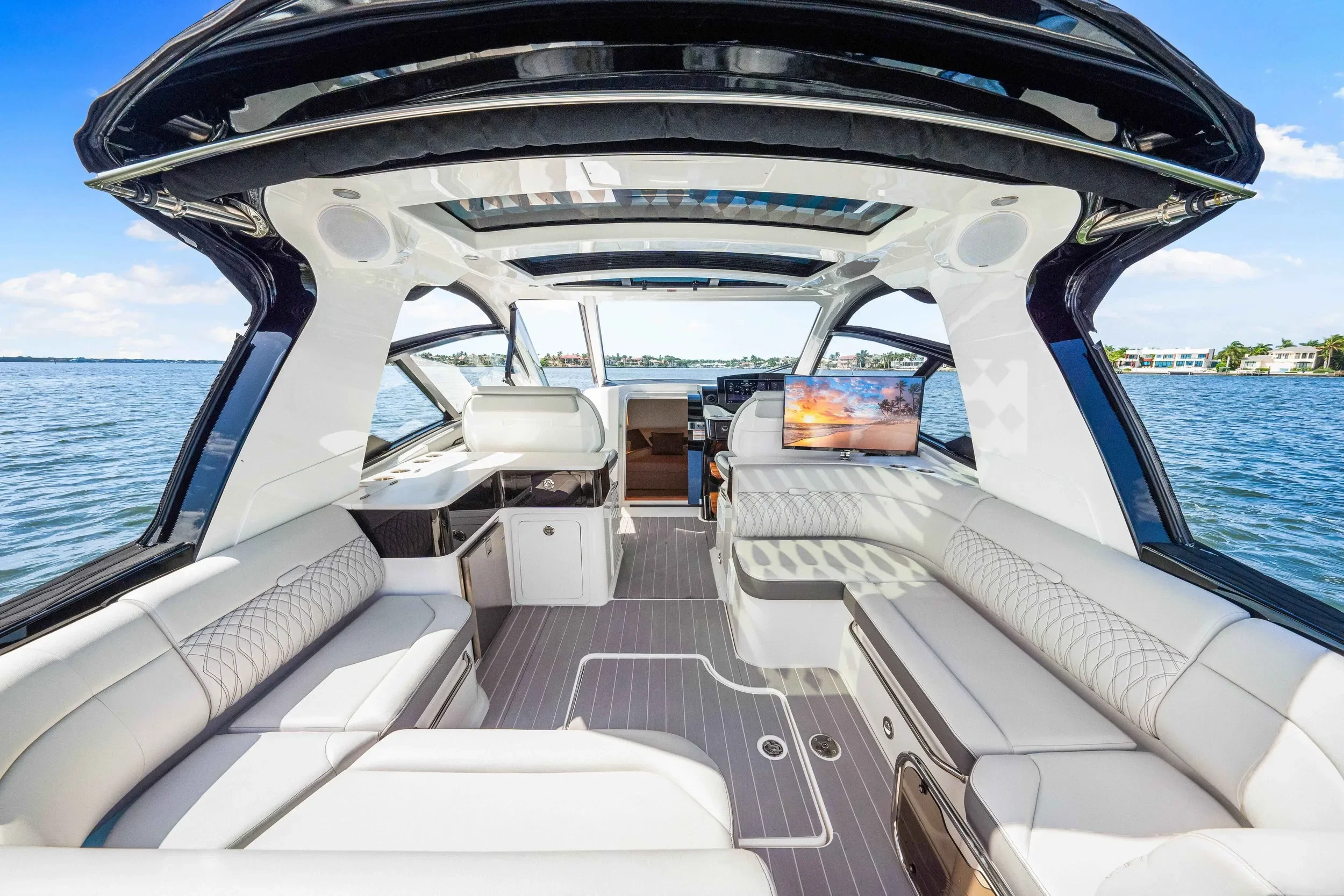 kokomo charters - 40' Golden Ticket - Interior view of a luxurious yacht with white leather seating, a flat-screen TV displaying a sunset, and large windows showing water and shoreline outside.