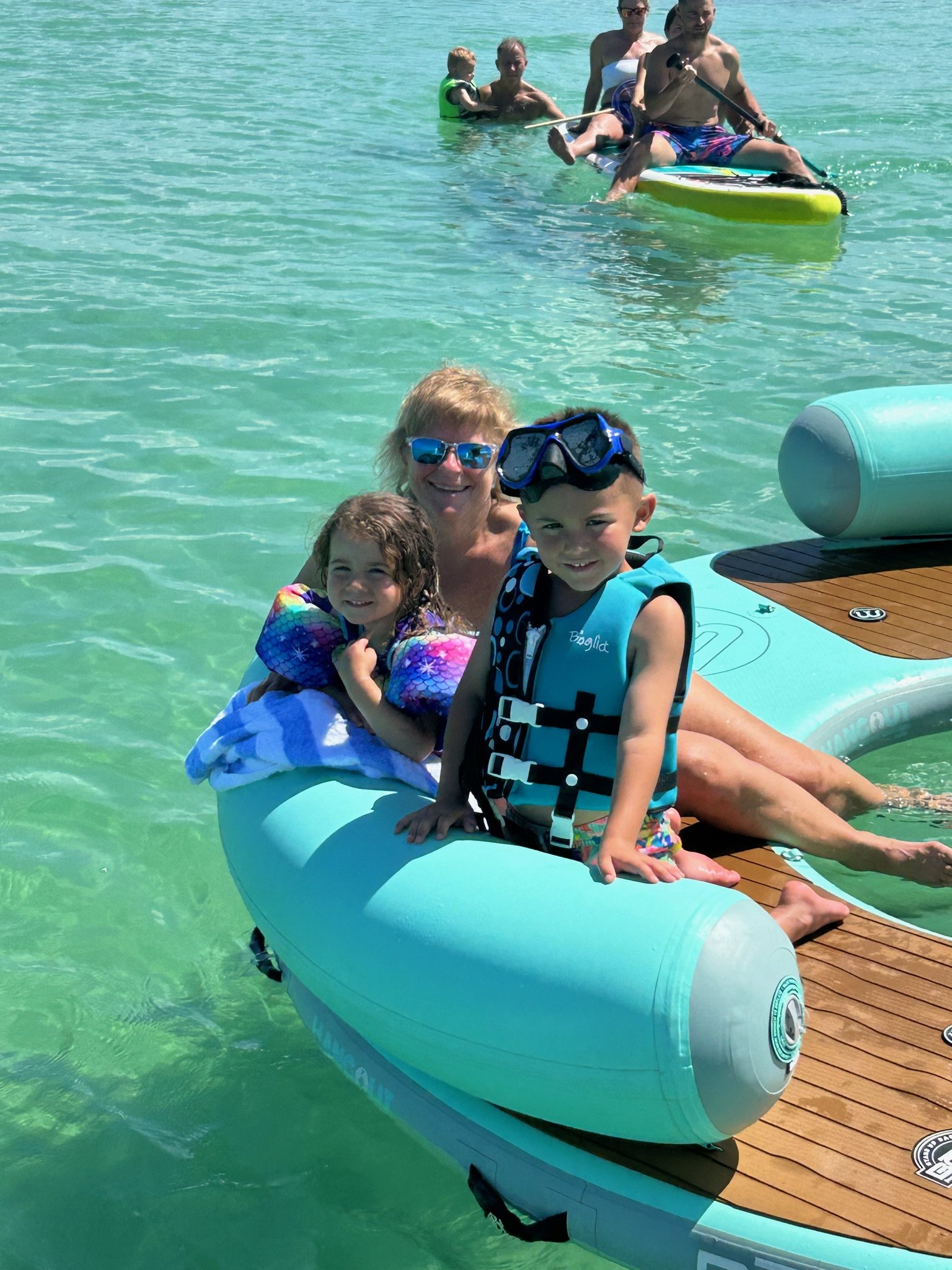 Children wearing life jackets on a floating platform with adults paddleboarding in the background, enjoying a sunny day on clear water.
