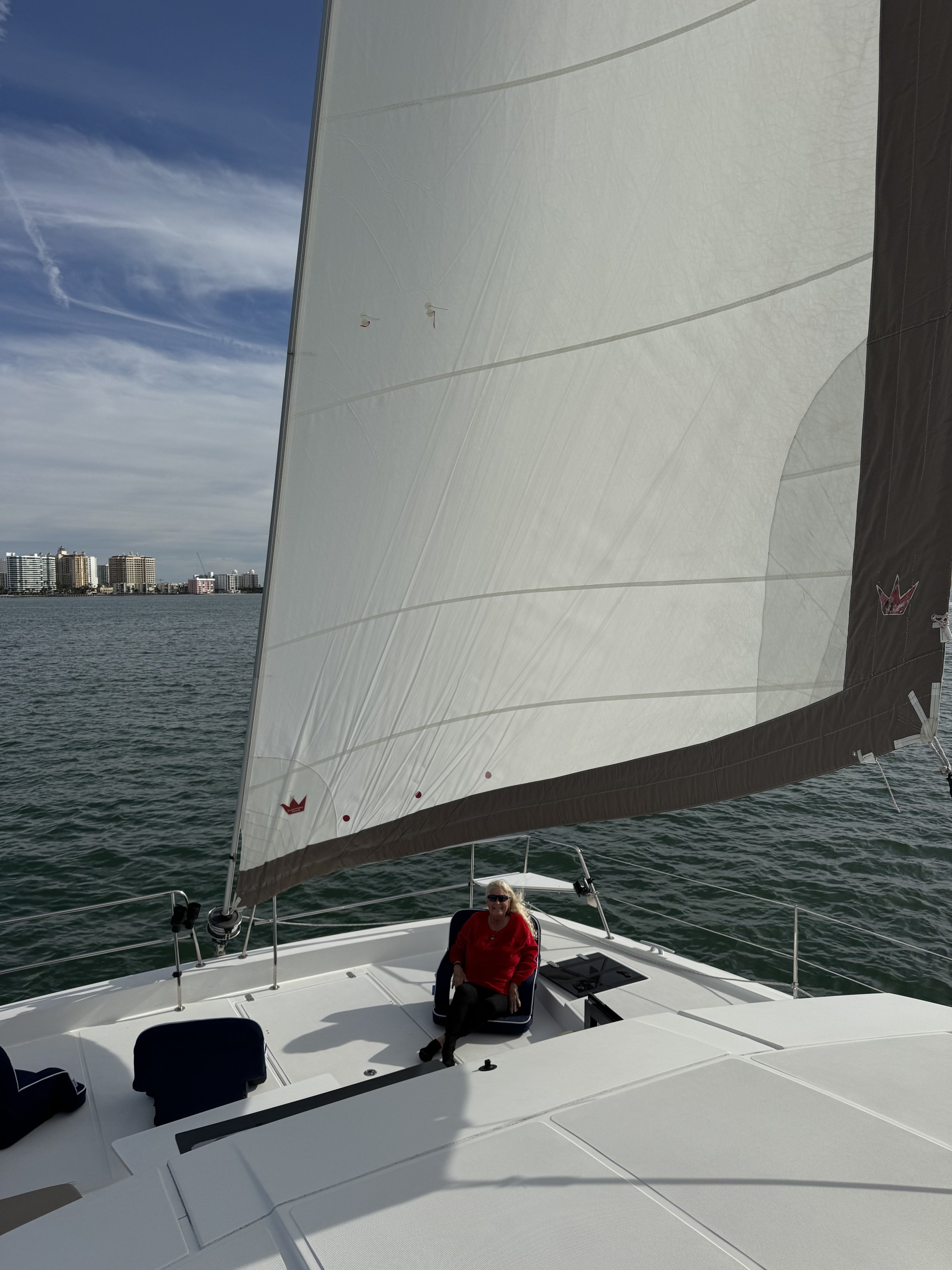 Person sitting on the deck of a sailboat with a large sail, in open water with buildings visible in the background.