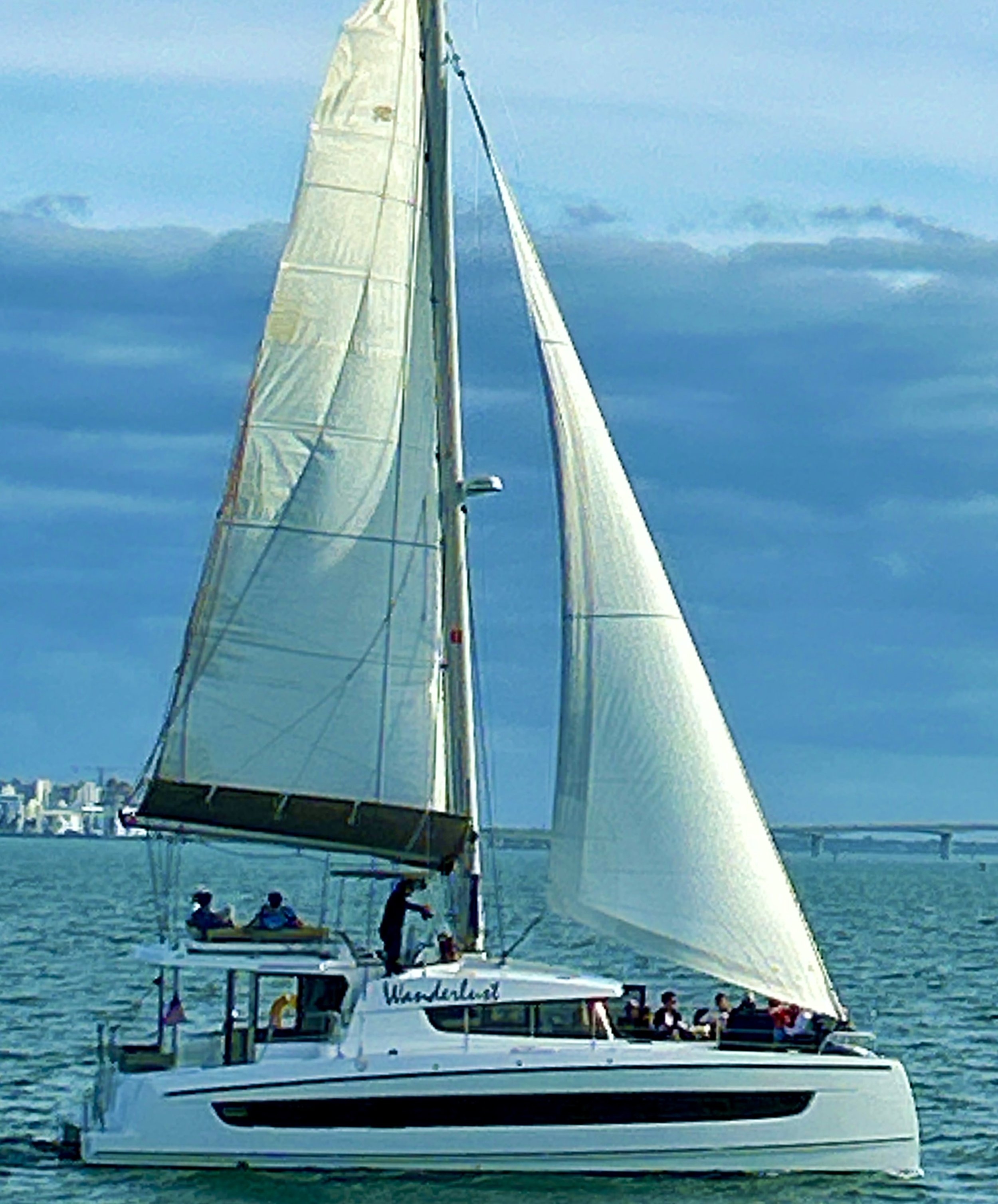 A catamaran sailboat on the water with several people on board, city skyline in the background.