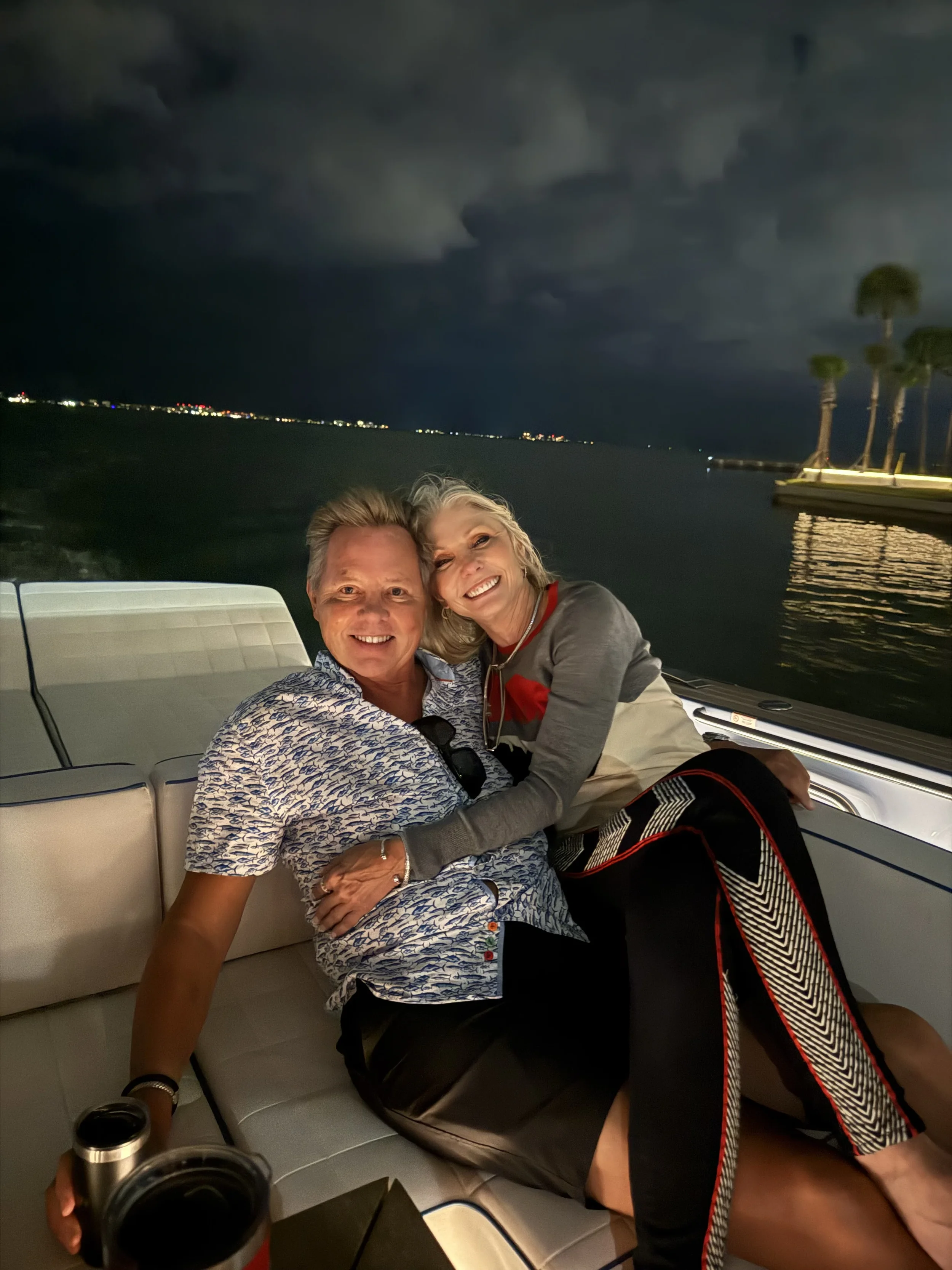 Two smiling people, a man and a woman, sitting close together on a boat at night with a dark sky and city lights in the background.