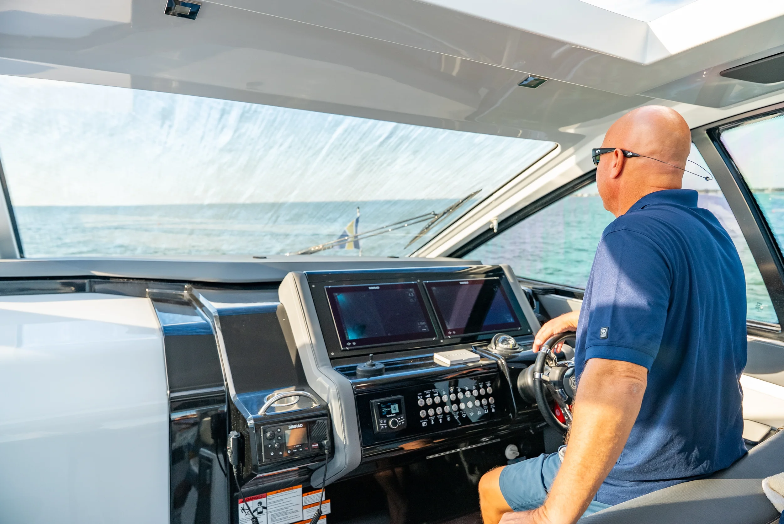 Kokomo charters - Sarasota private yacht charter - A man with a bald head wearing sunglasses and a blue shirt sitting at the controls of a yacht, steering through open water with a clear sky.
