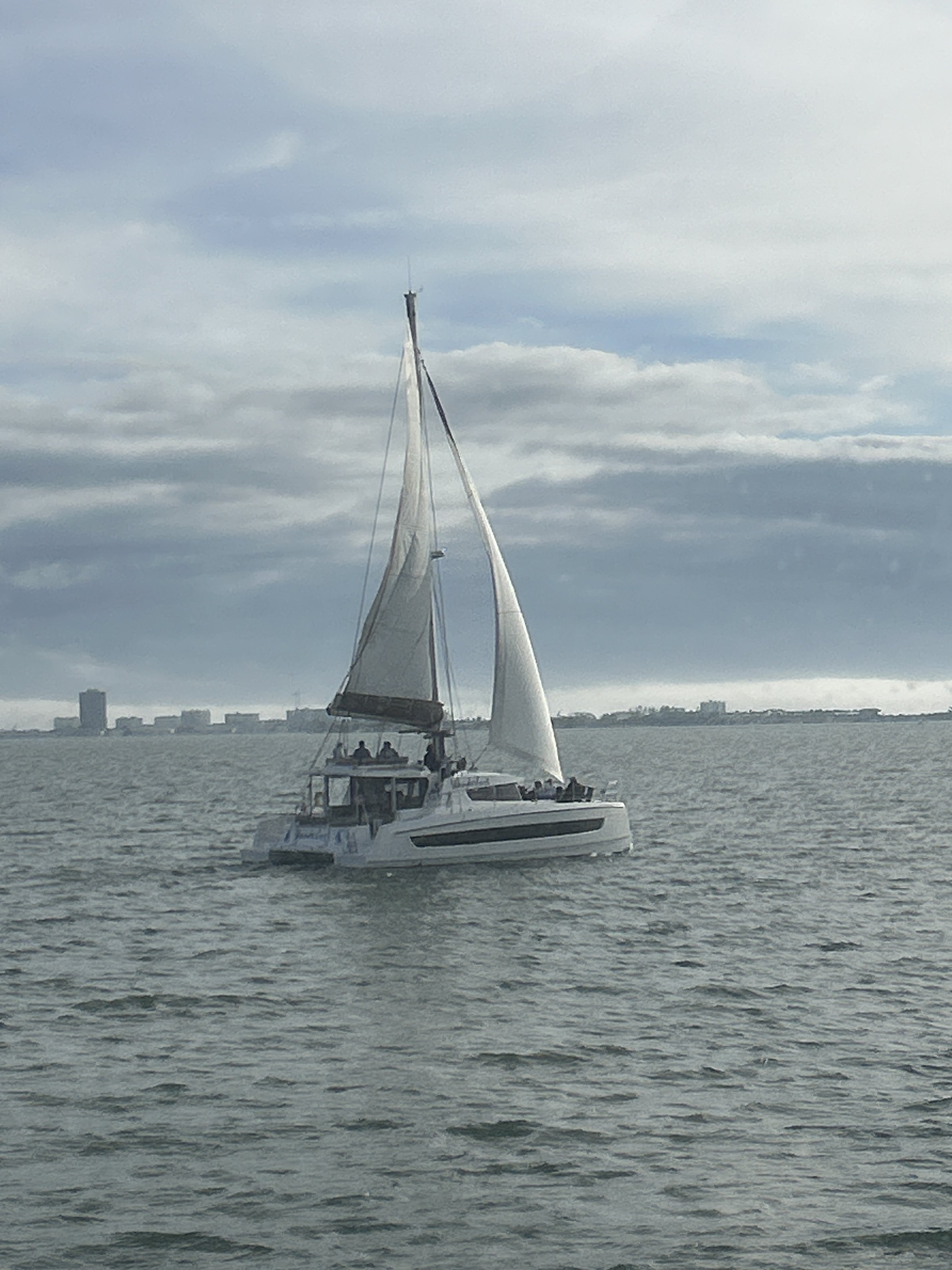 Sailboat on a calm sea with overcast sky and city skyline in the background.
