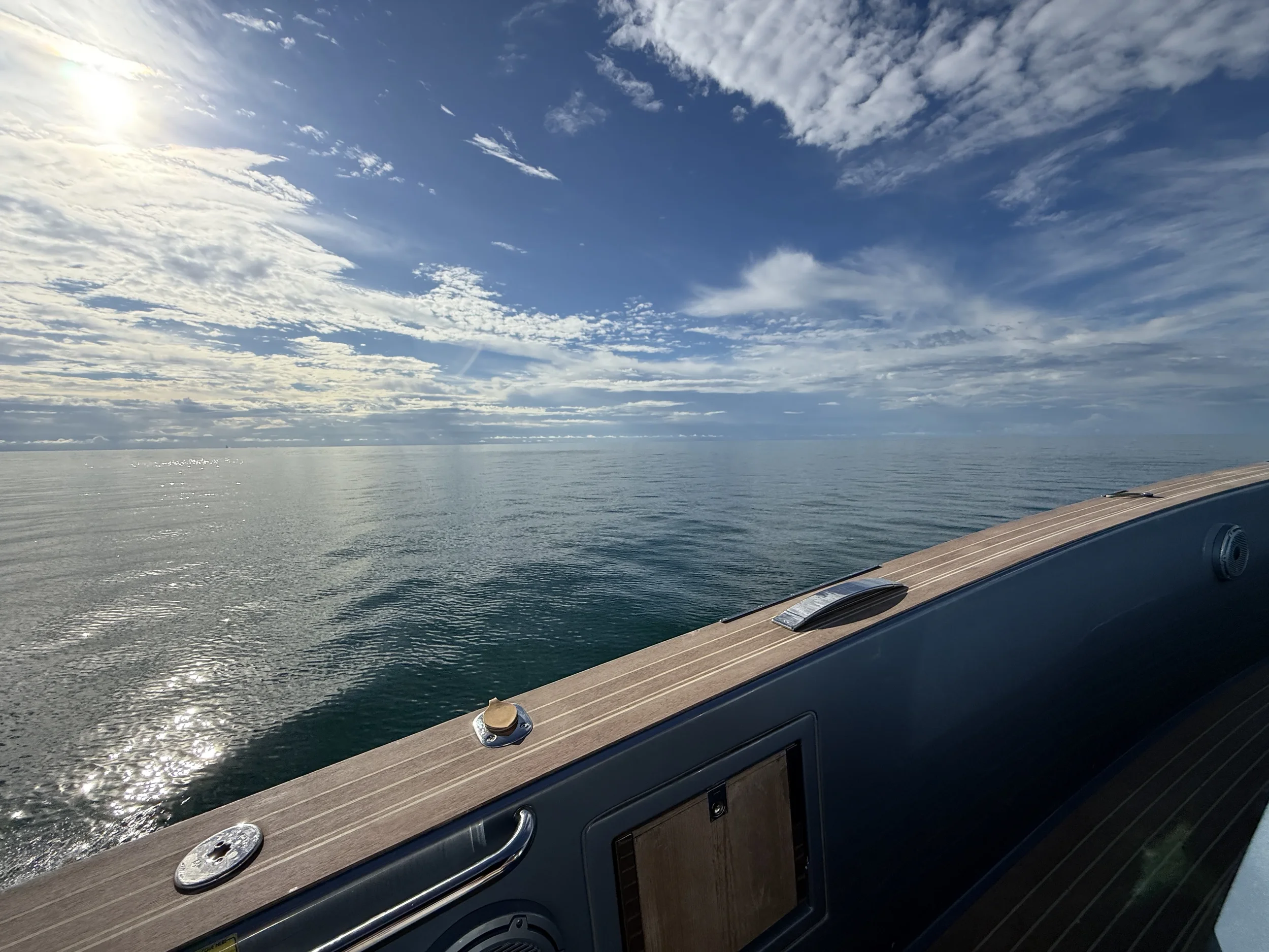 View of open ocean with partly cloudy sky and sunlight reflecting on the water seen from a boat deck with a wood panel railing.