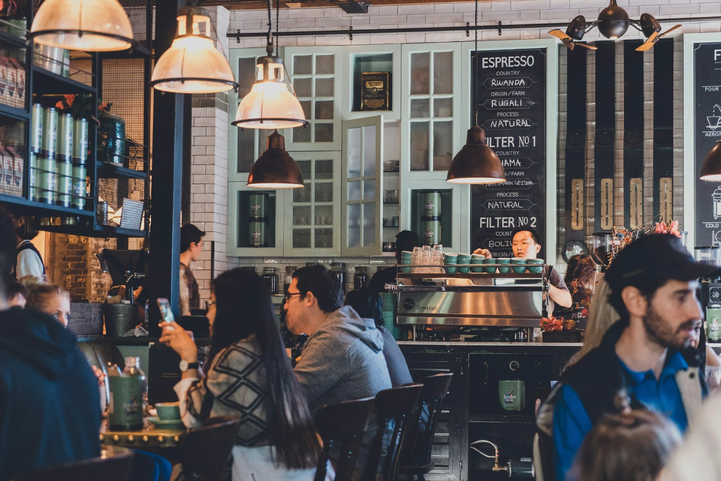 Inside a busy coffee shop with customers sitting at tables, a barista working behind the counter, industrial-style hanging lights, chalkboard menu, and shelves with coffee supplies.