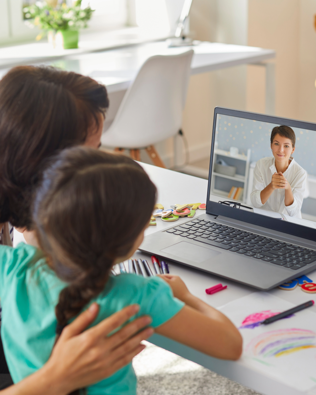 A woman and a young girl are watching a video call on a laptop, with the woman having her arm around the girl in a supportive gesture.