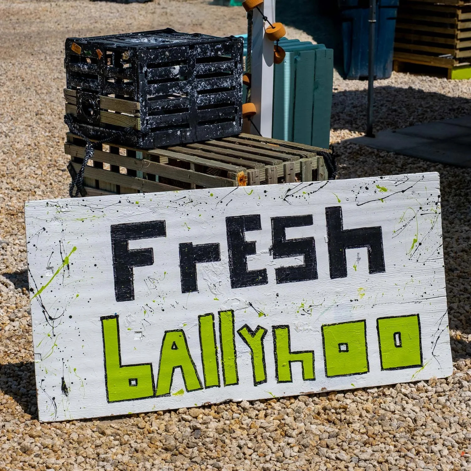Handmade sign with black and green lettering on white painted wood, reading 'FRESH LAILYN-LO' with paint splatters, placed on gravel surface with a black and green wooden crate and small skateboard in the background.