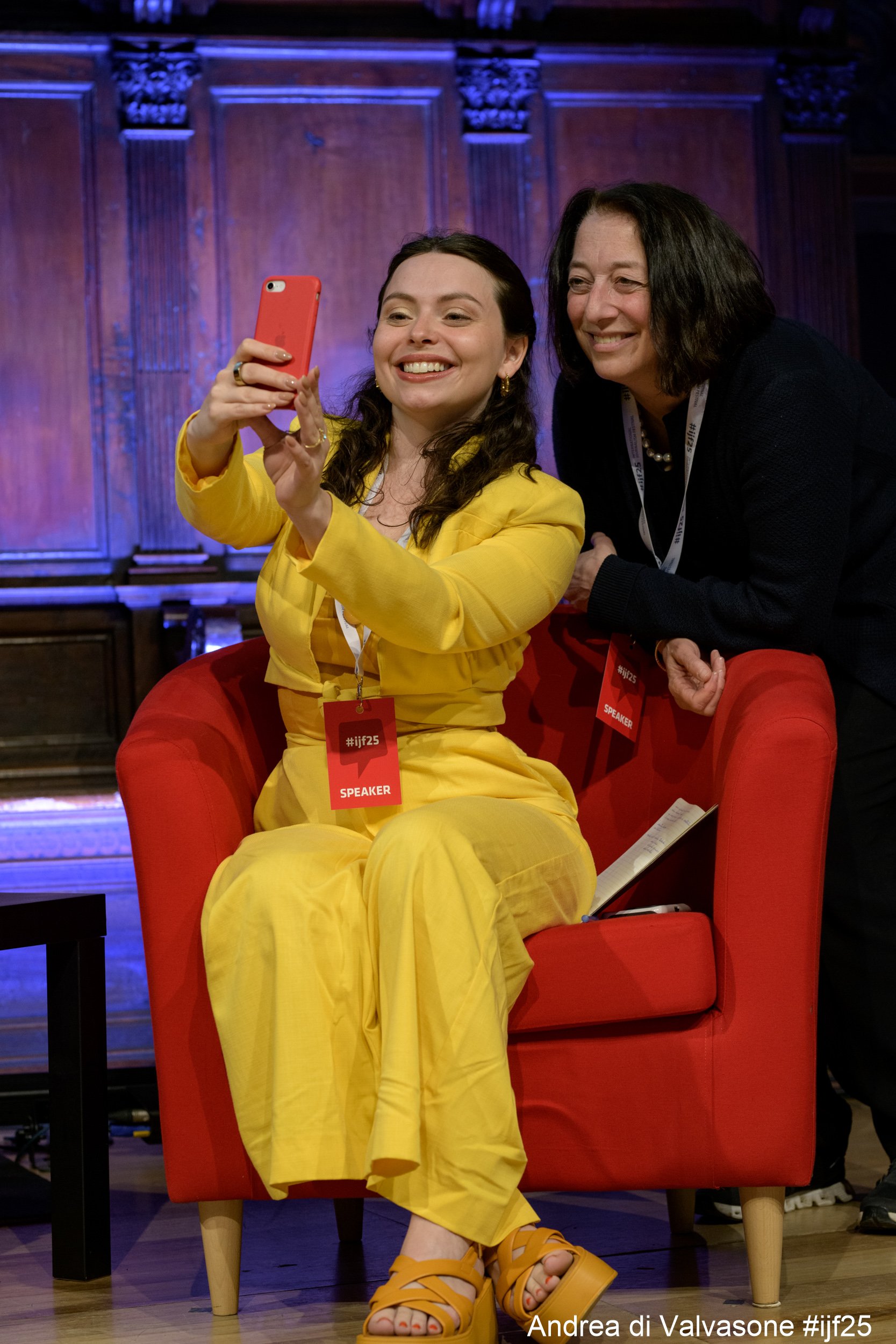 A woman in a yellow outfit taking a selfie with another woman in a black outfit at a conference or event.
