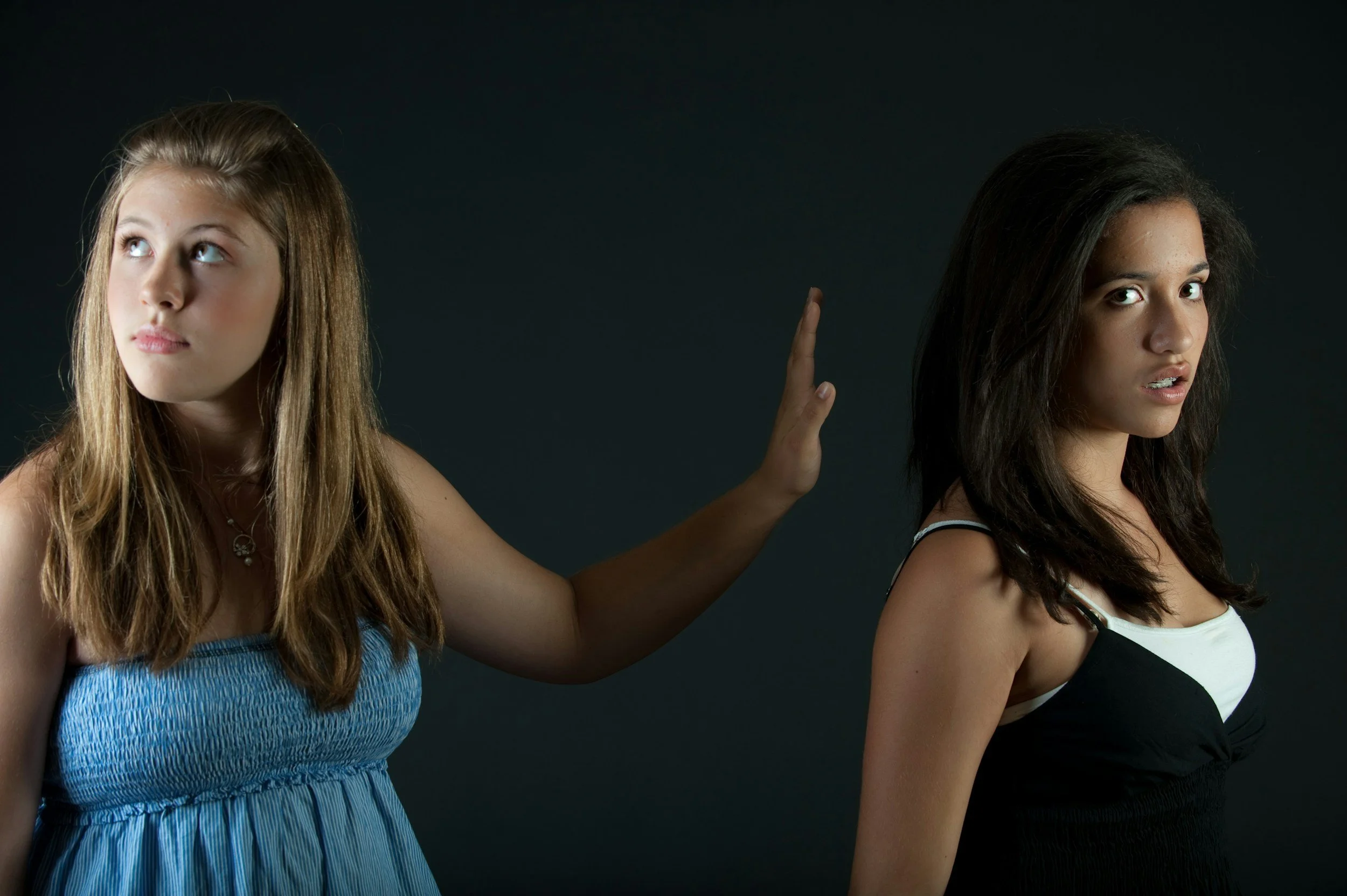 Dos mujeres jóvenes, una mirando de lado con la mano levantada y la otra mirando hacia la cámara, ambas sobre un fondo oscuro. Representa las dificultades en la comunicación, en las relaciones.