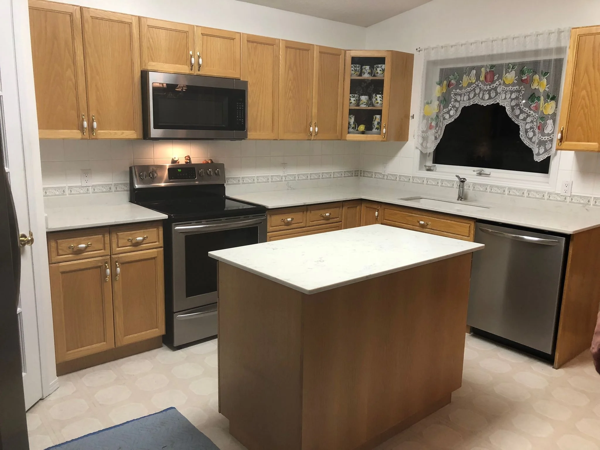 Kitchen with wooden cabinets, microwave, stove, dishwasher, and a kitchen island with a quartz countertop. There is a window with lace curtains over the sink.