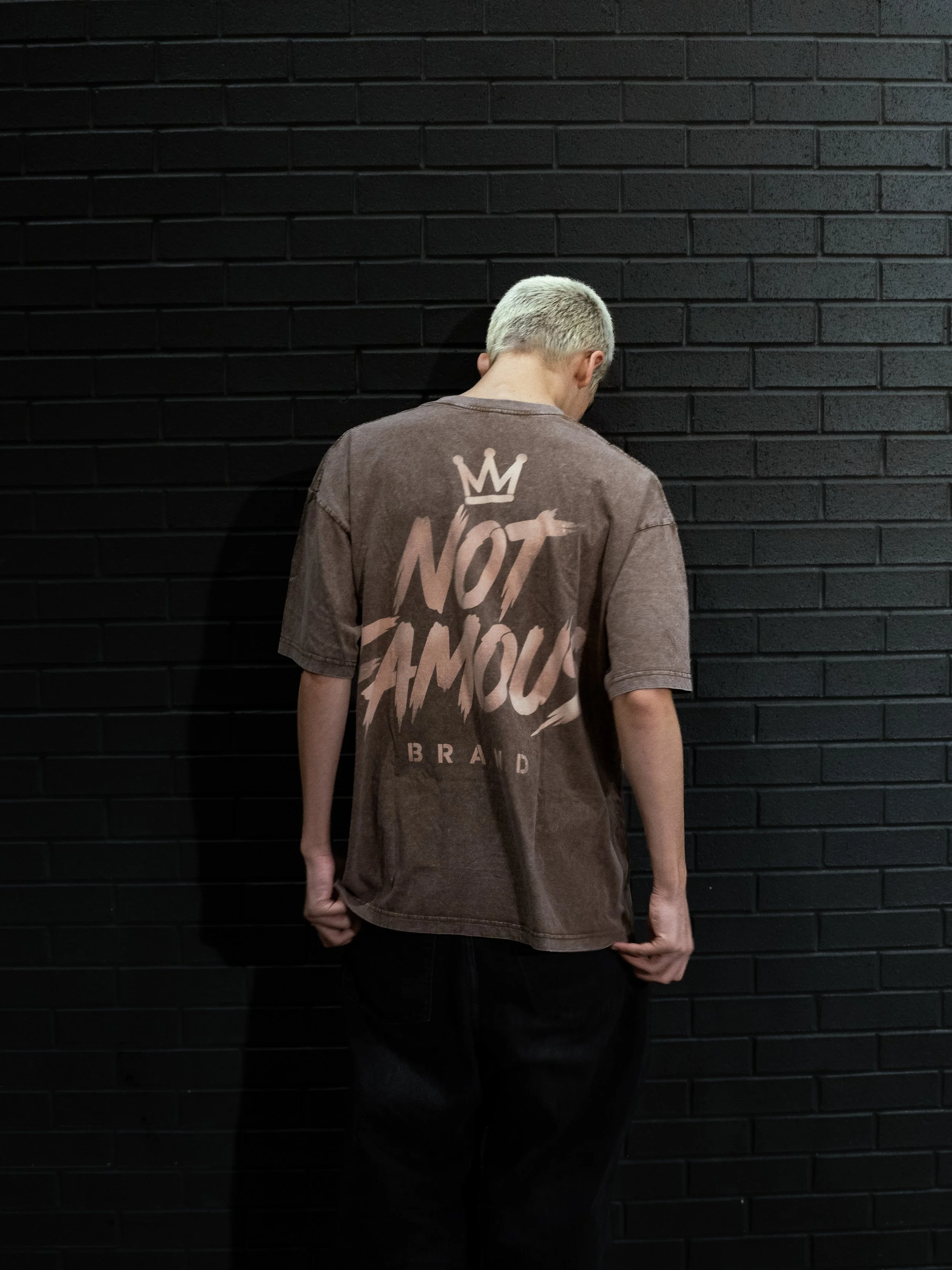 A person with short blond hair facing a black brick wall, wearing a brown T-shirt with the words 'NOT THE FAMOUS' on the back and a crown symbol above the text.