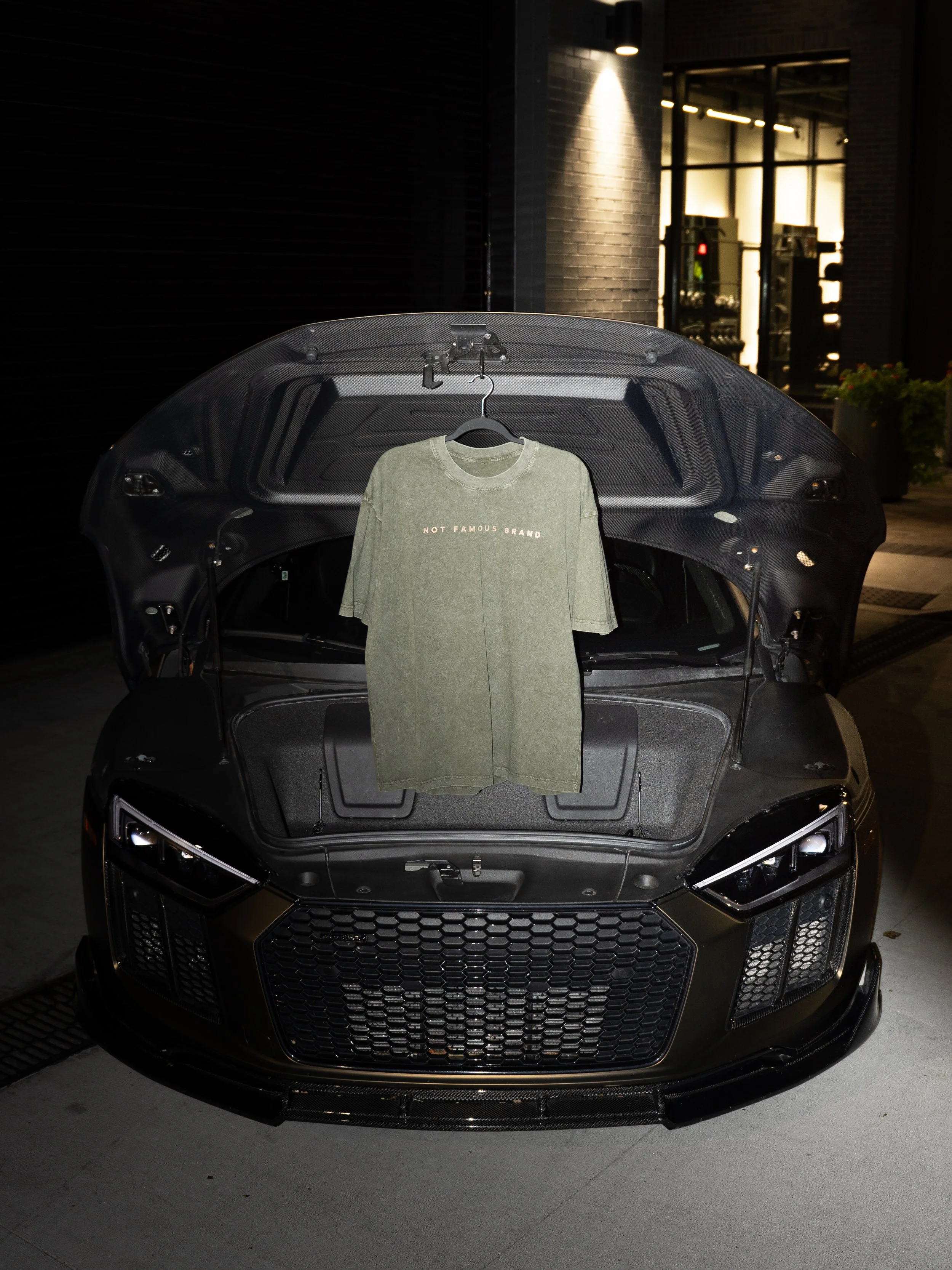 A black car with its hood open, showing the engine compartment at night, with a grey T-shirt hanging from a hanger inside the car that reads 'NOT FAMOUS BRAND'.