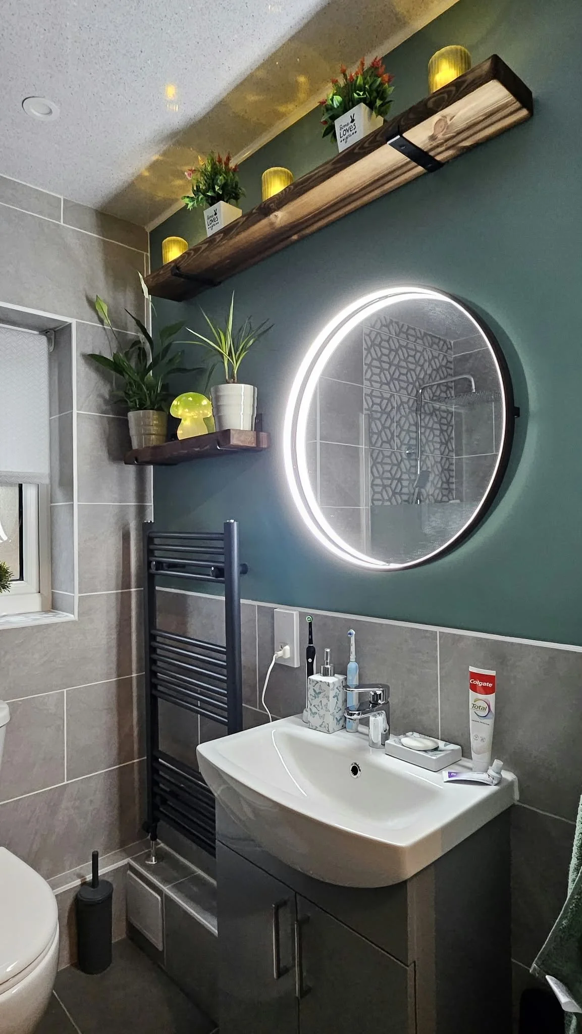 Modern bathroom with illuminated round mirror, wall shelves with plants and lights, white sink vanity, tiled walls, and black towel radiator