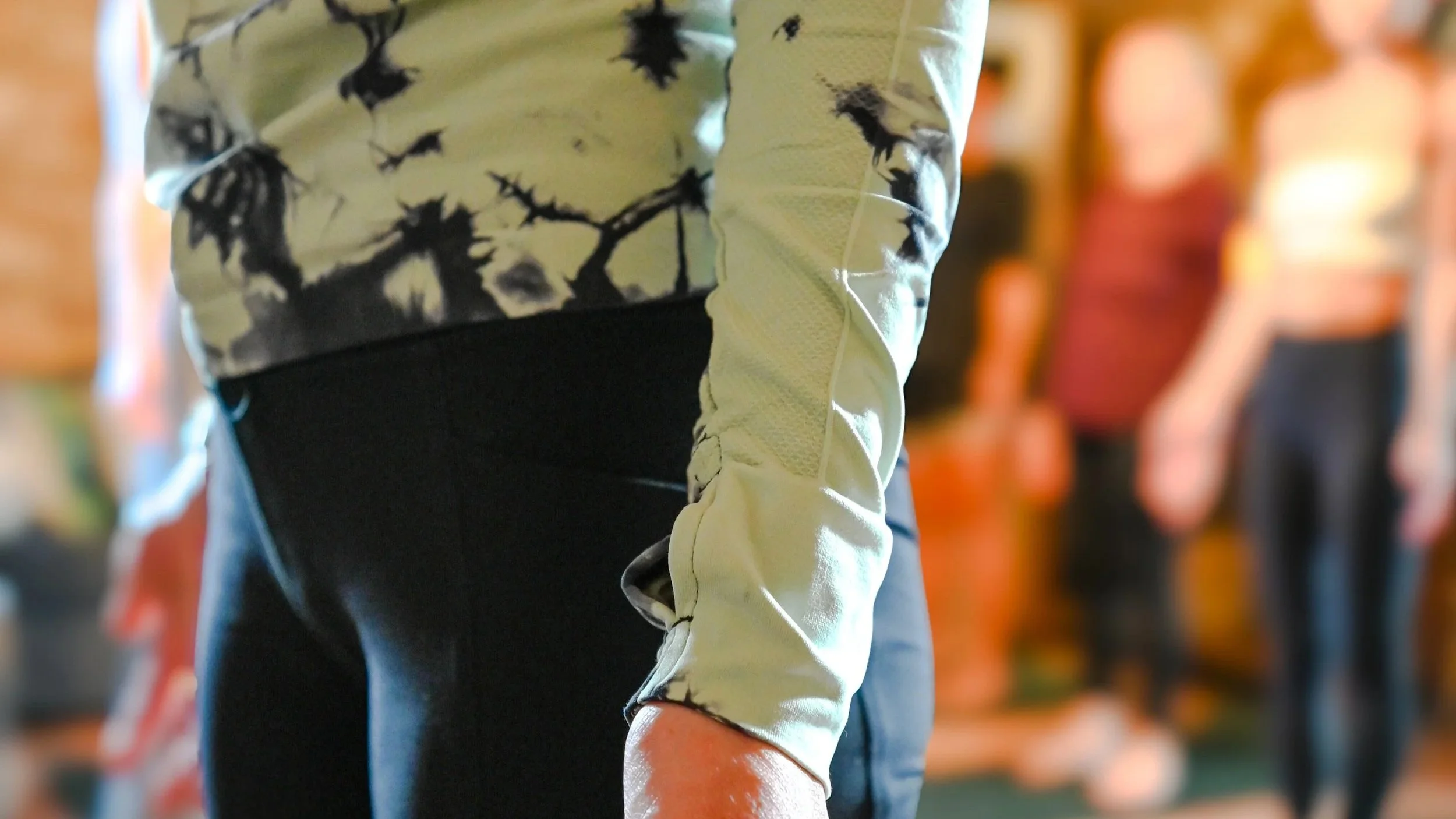 A person wearing a light-colored long sleeve shirt with black tie-dye patterns, standing next to other people in an indoor setting, possibly a gym or fitness class.